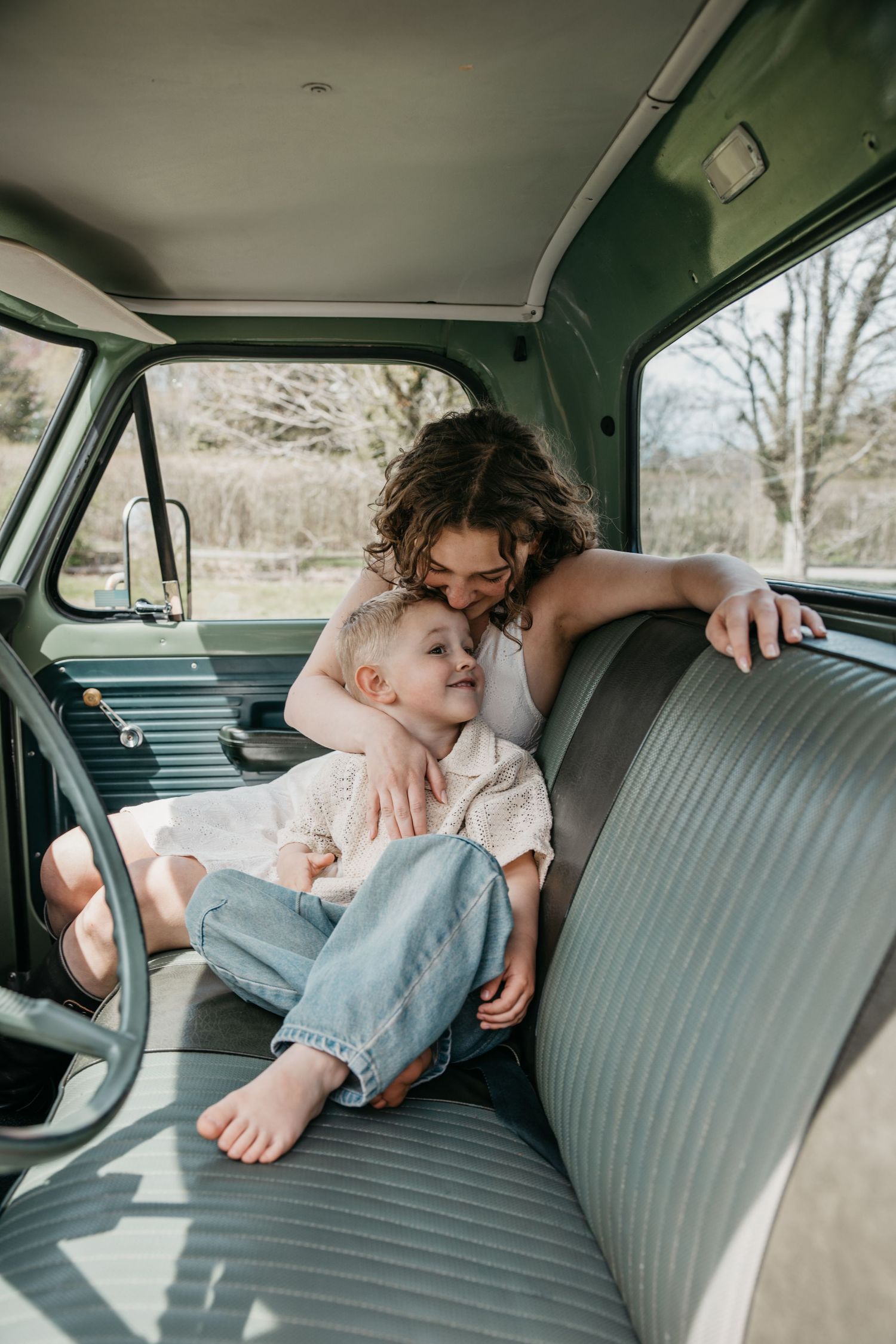 A mother tenderly embraces her young child on the back seat of a vintage green truck cab.