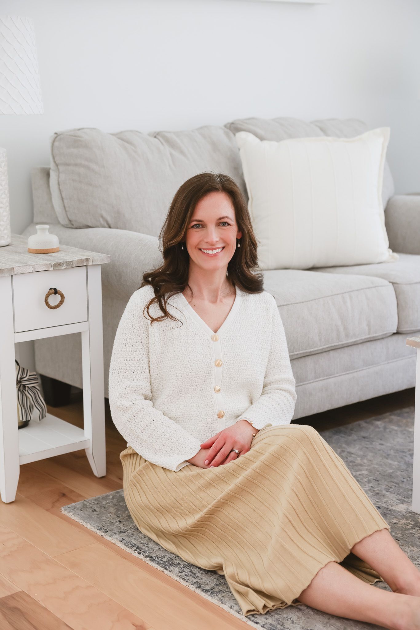 A smiling brunette woman sits on a rug in a bright living room wearing a white cardigan and gold pleated skirt.