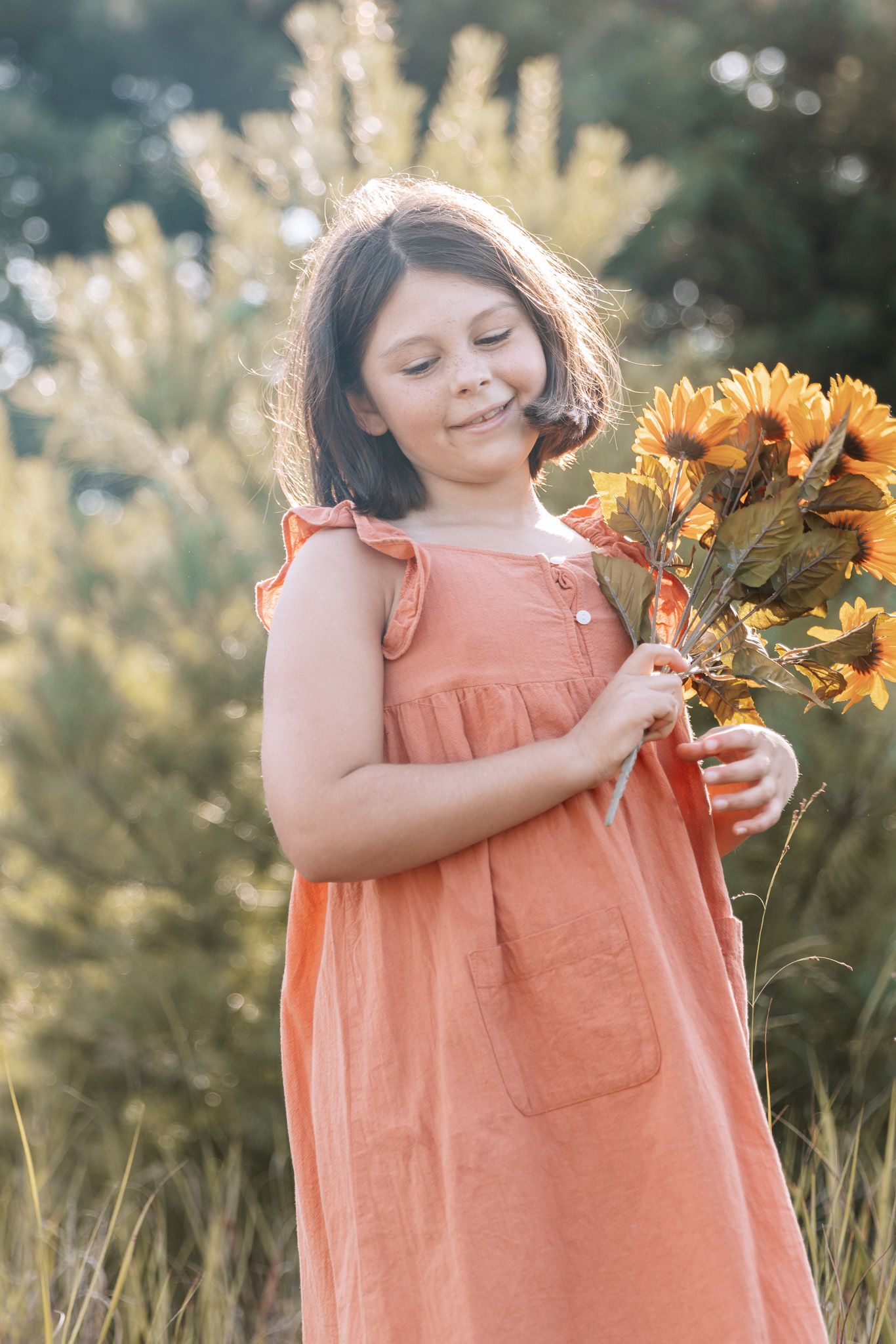 A child in a peachy-orange dress admires bright sunflowers in a natural outdoor setting.