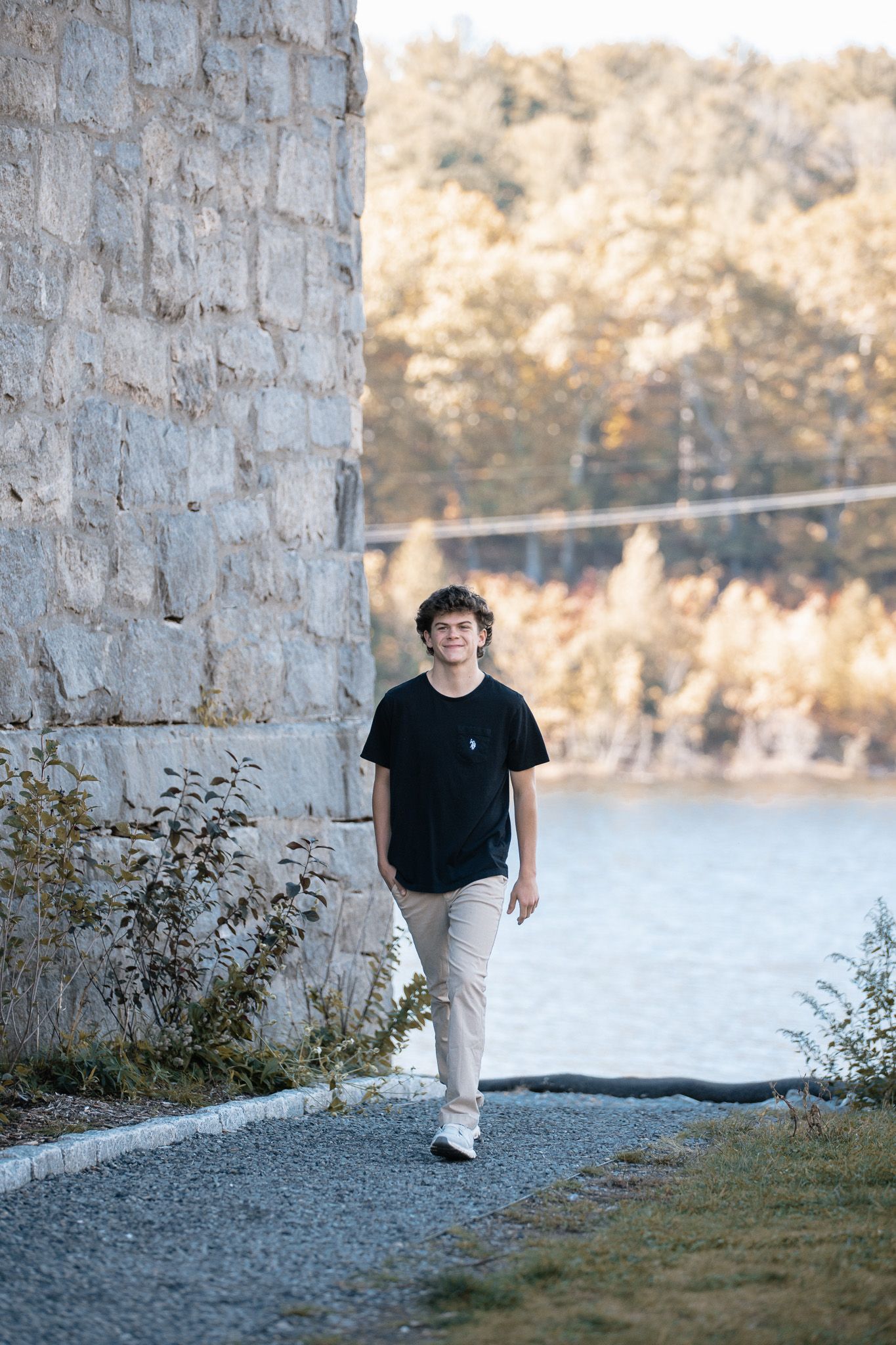 Teen in black t-shirt walking near stone wall with water view in background.