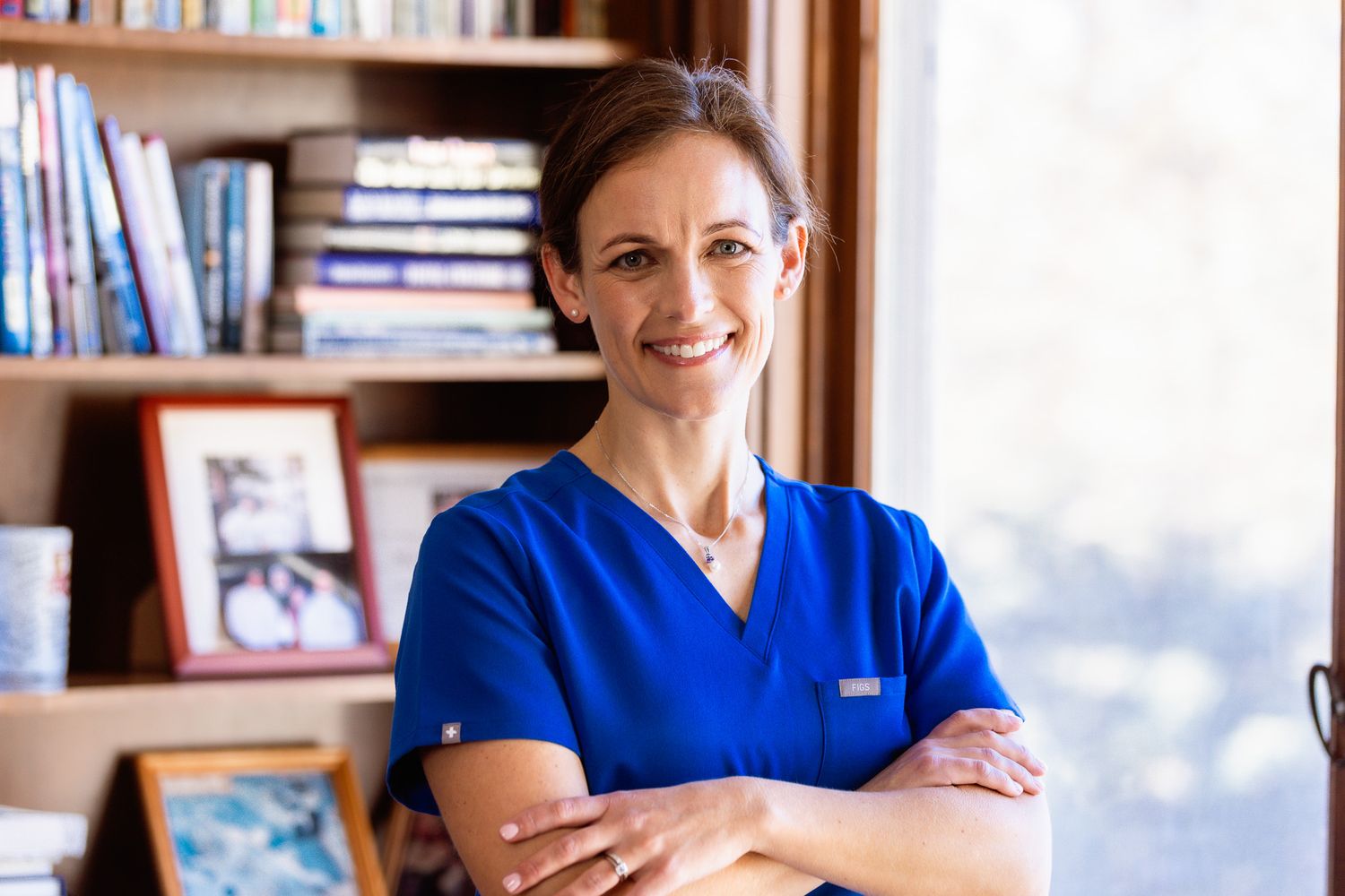 Healthcare professional in blue scrubs stands in front of a bookshelf in an office setting.
