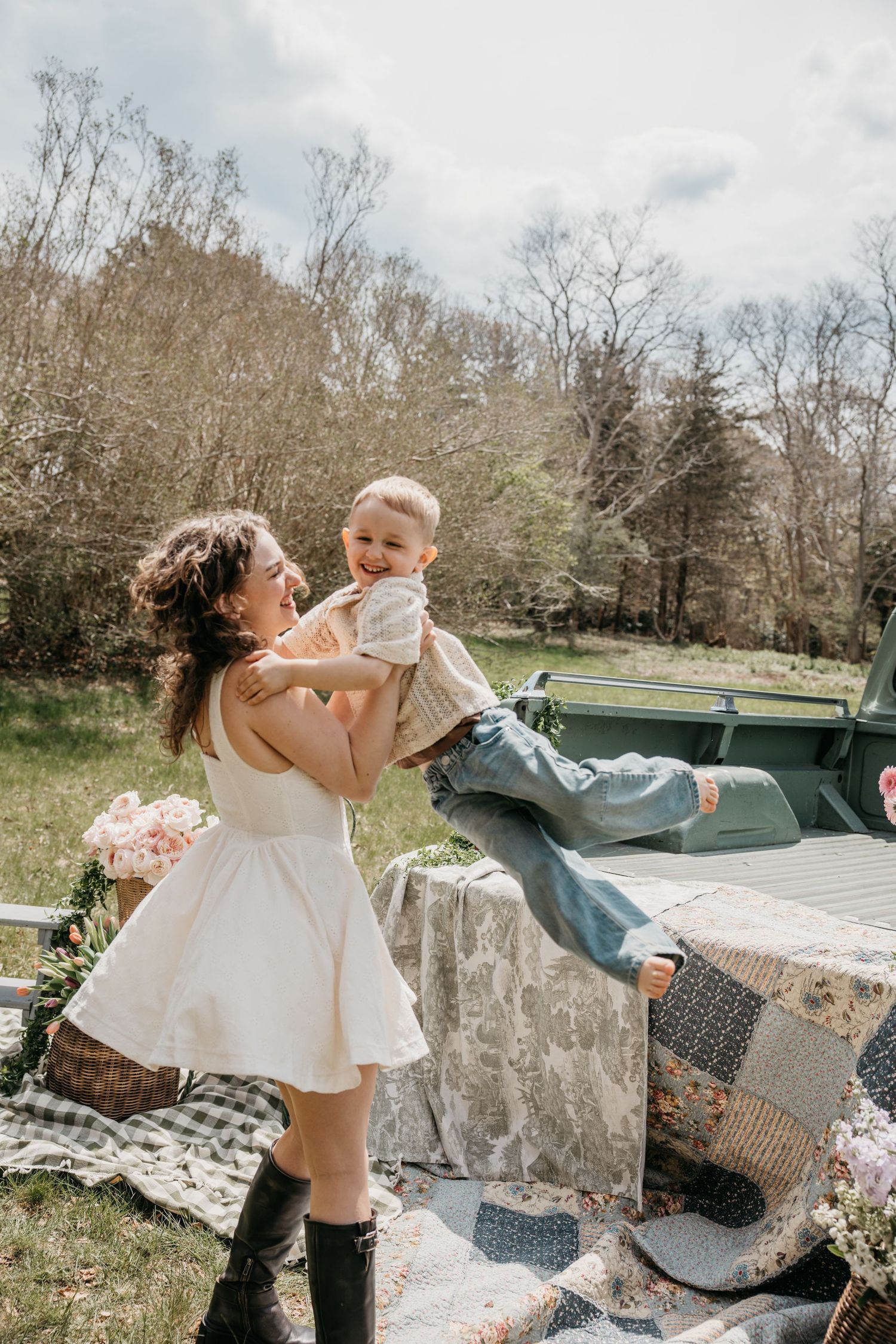 A joyful mother in a white dress lifts a laughing toddler during an outdoor picnic with a vintage truck backdrop.