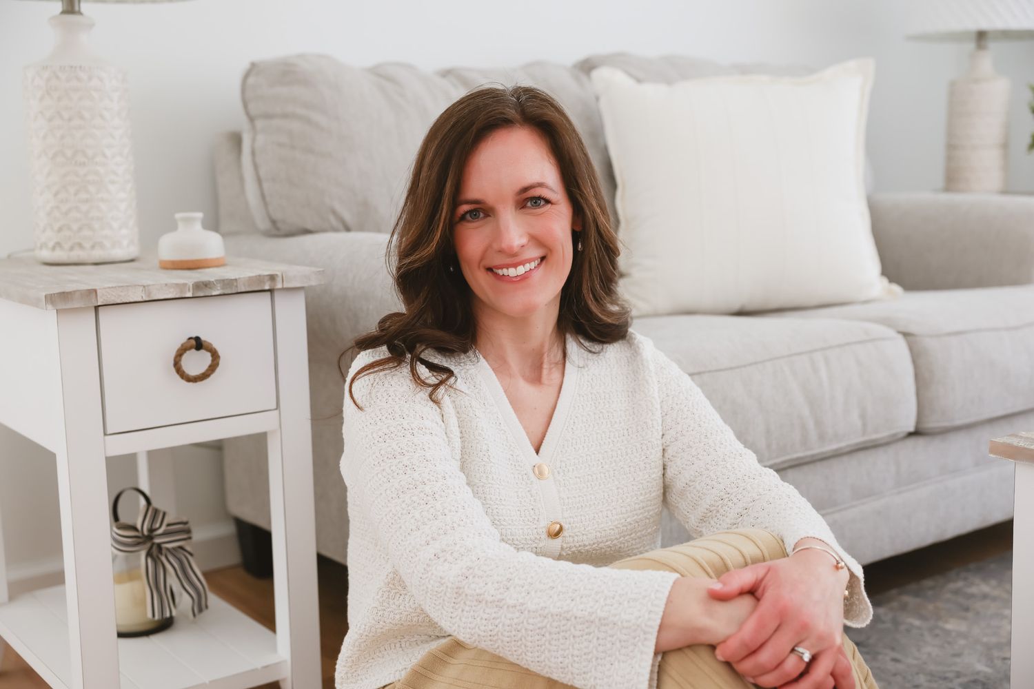 A smiling woman in a white cardigan sits cross-legged in a bright, modern living room with neutral decor.