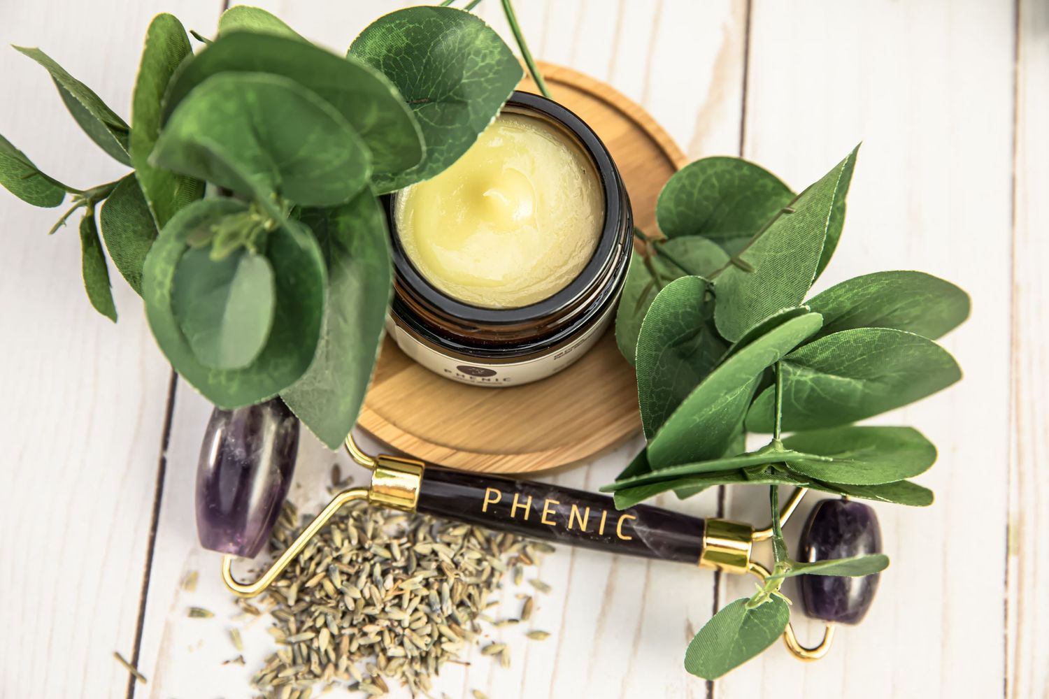Natural skincare product with green leaves and dried herbs on white wooden surface.