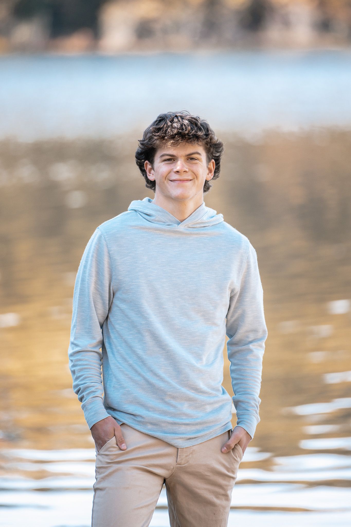 Teen in light gray hoodie poses by water during golden hour photoshoot.