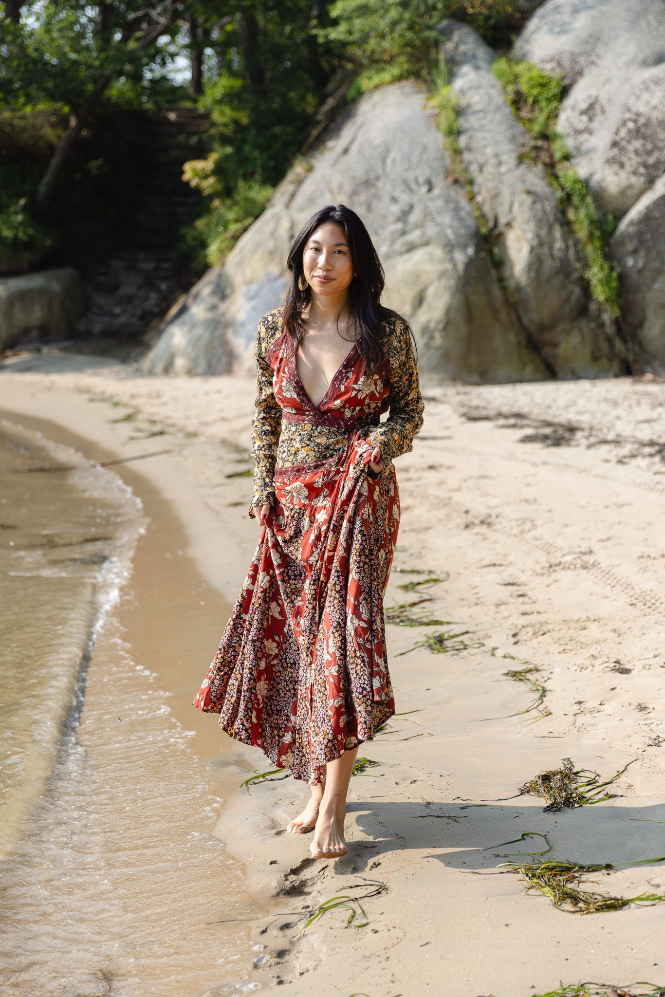 Woman in flowing red floral dress walks barefoot along a rocky beach.