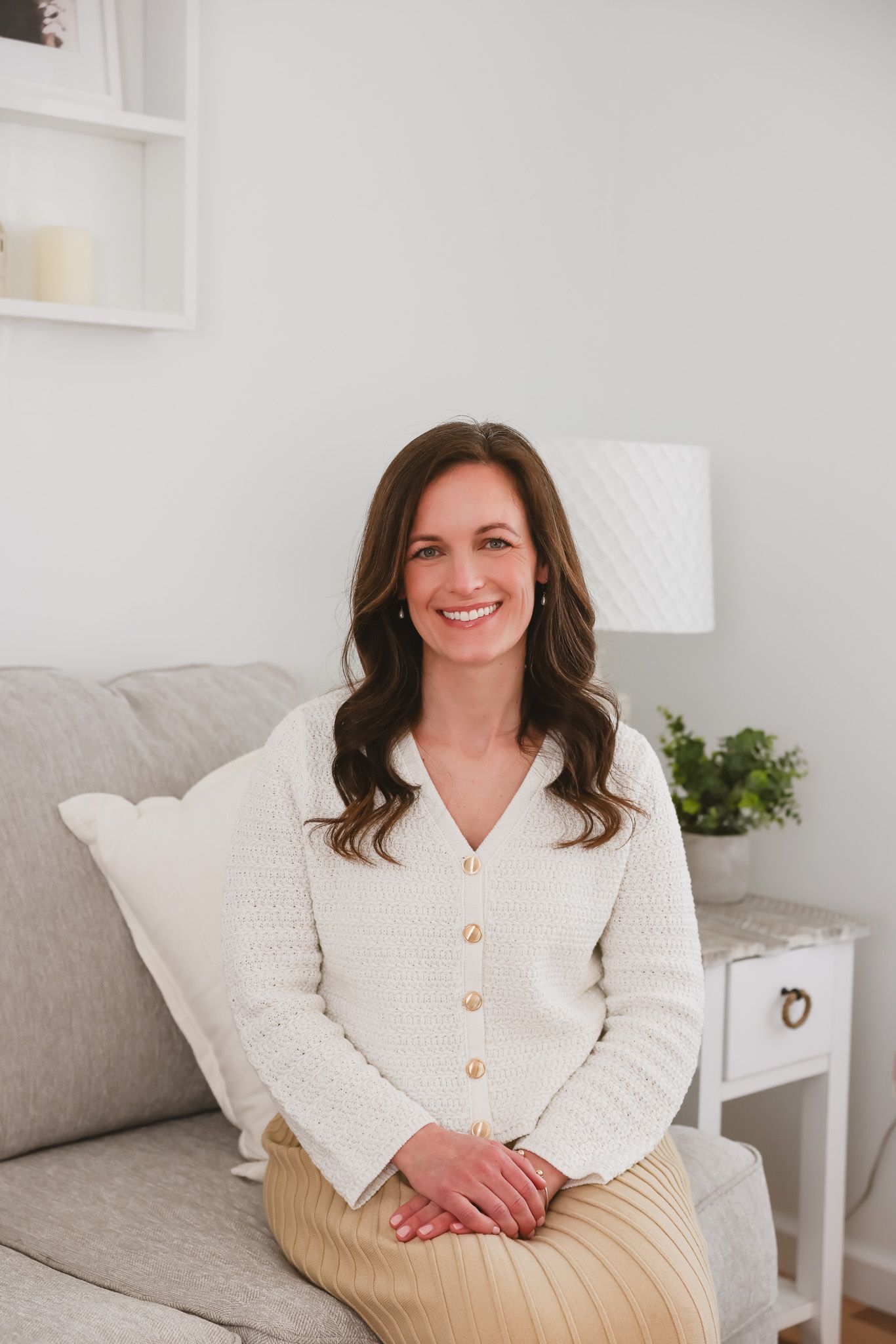 Smiling woman in white cardigan and gold skirt sitting on a gray sofa in a bright, modern living room.