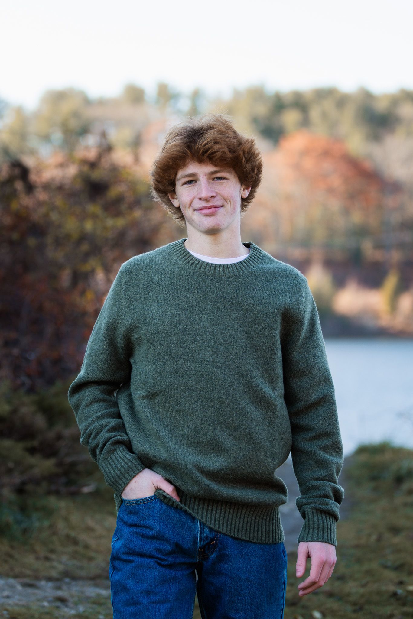 Figure in a cozy green knit sweater stands by a lake during fall season.