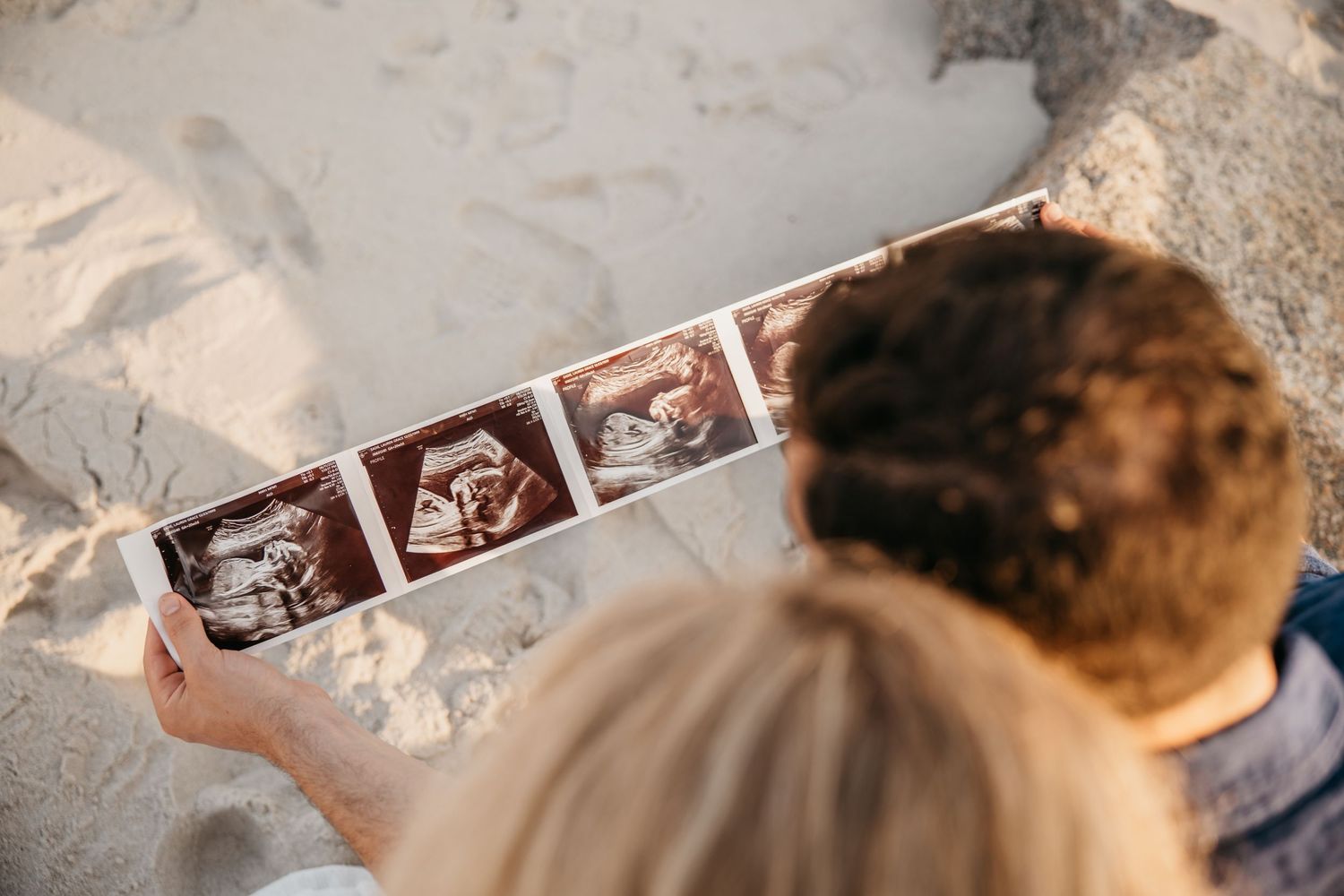 Couple sitting on sandy beach looking at ultrasound photos, pregnancy announcement moment captured from above.