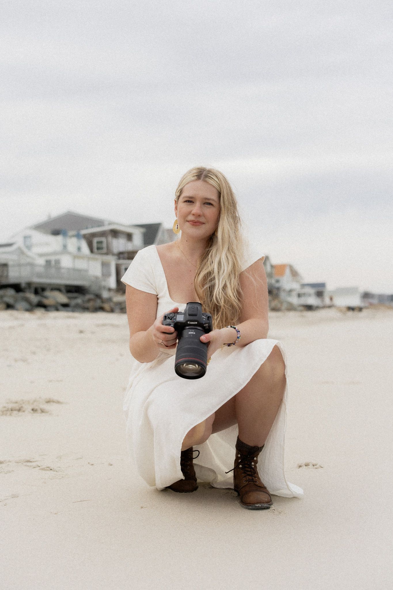 Photographer in white dress crouches on sandy beach with camera while coastal homes line the shore in background.