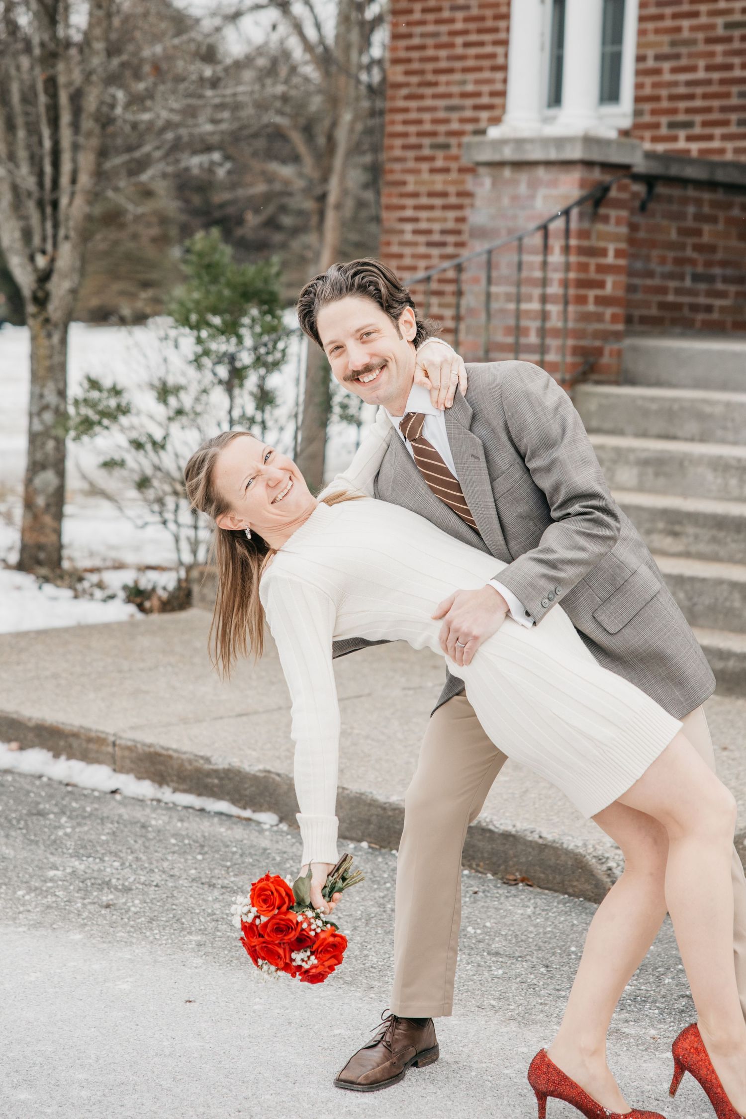 Romantic couple in formal wear dips and poses playfully outside brick building.
