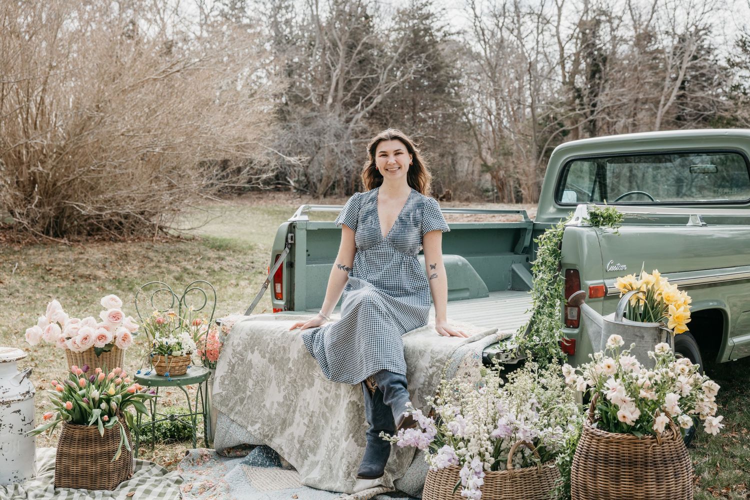 Woman in gray dress and hat sits on vintage green truck surrounded by spring flowers in a rustic outdoor setting.