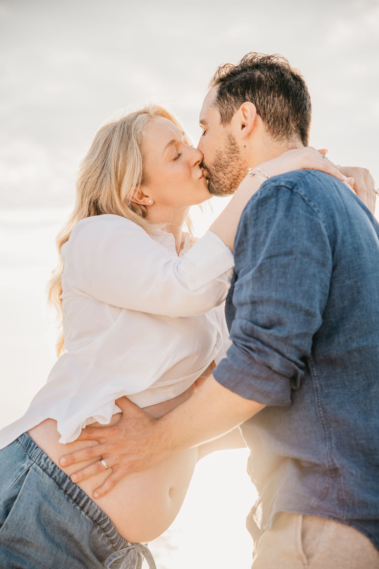 Pregnant couple sharing a romantic kiss on the beach at golden hour, woman in white shirt and jeans.