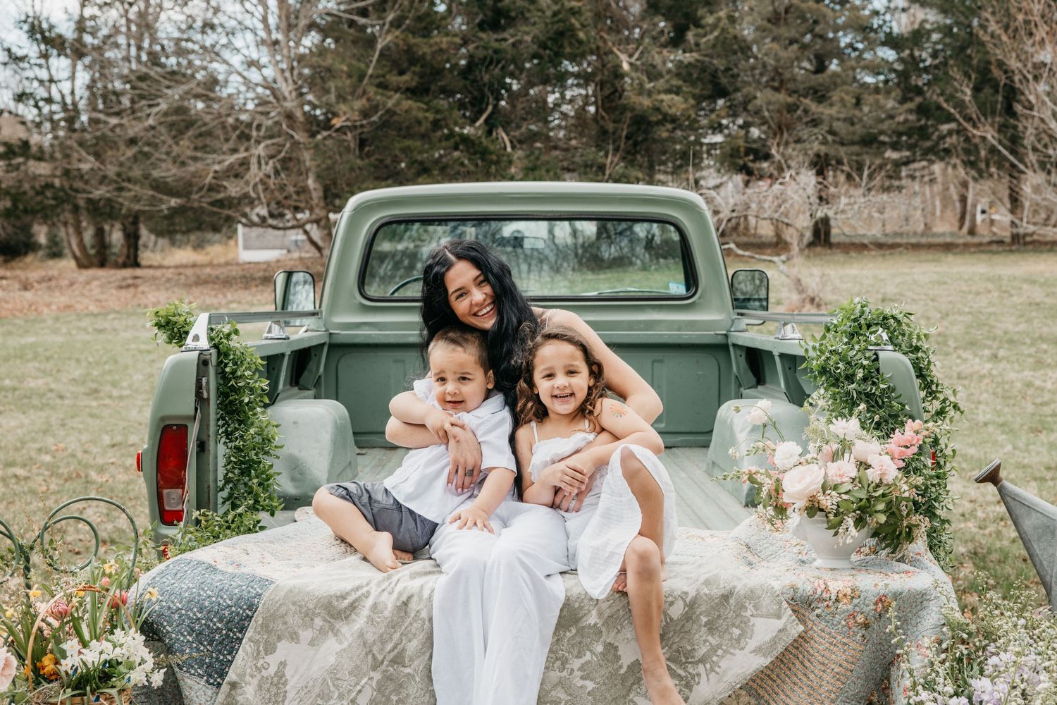 Woman in white dress sits with two young children in vintage green truck bed surrounded by floral decor outdoors.