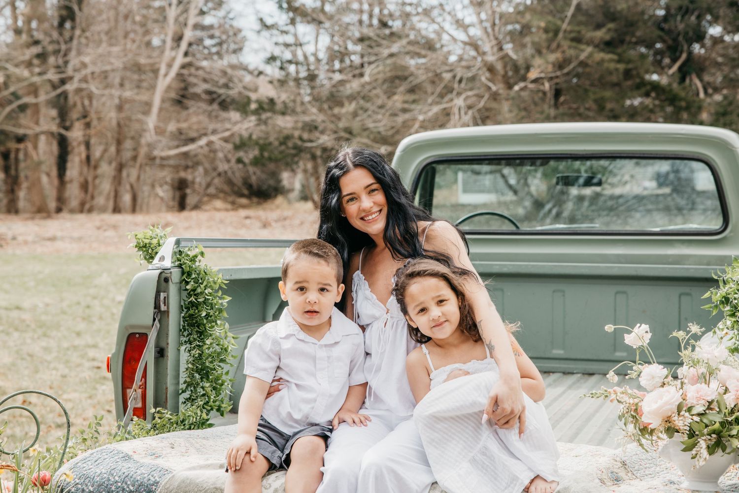 A woman in white dress sits with two young children on a vintage green truck bed surrounded by spring florals.
