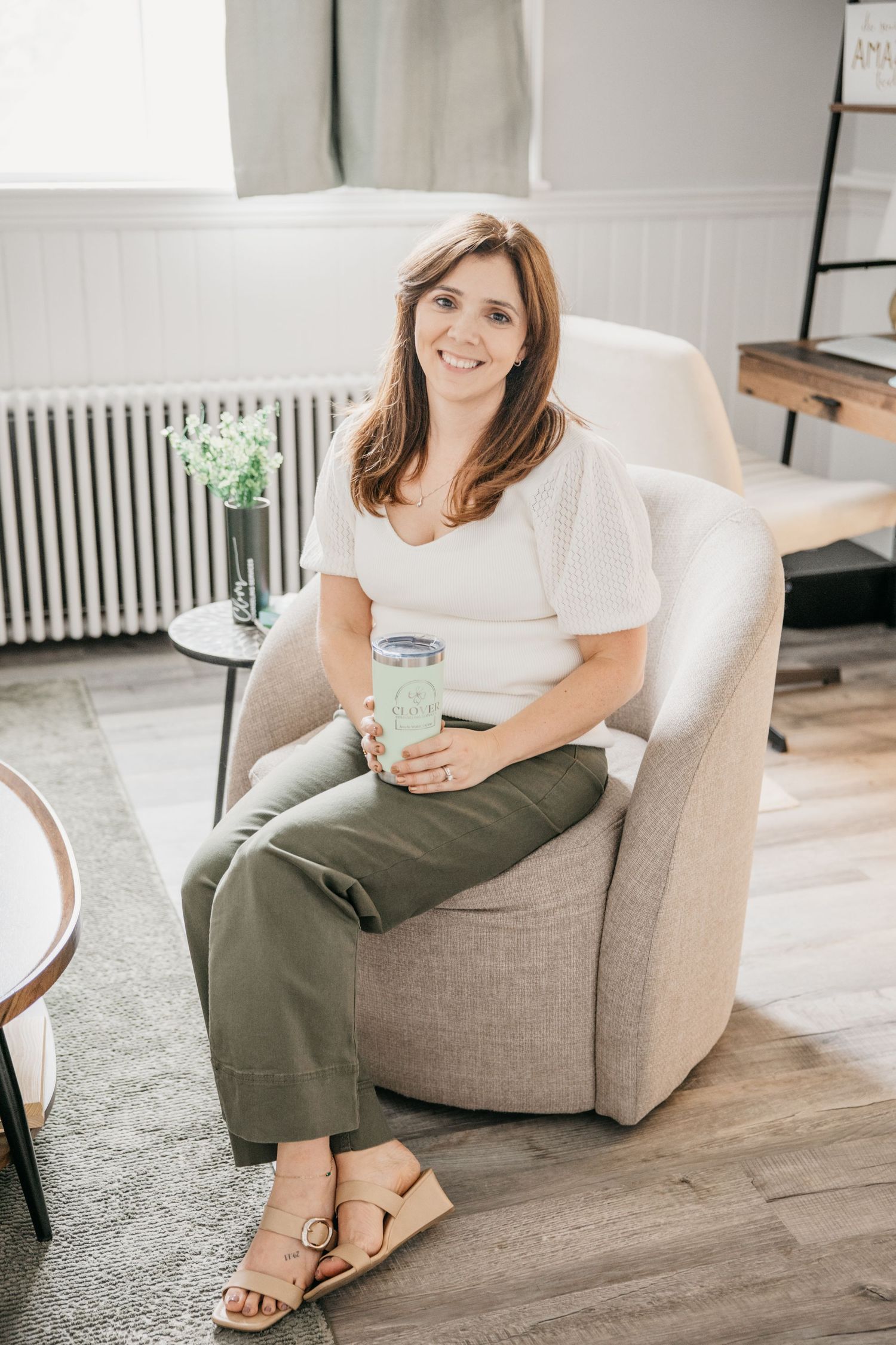 Smiling brunette woman sits in gray chair holding green tumbler, wearing white top and olive pants.