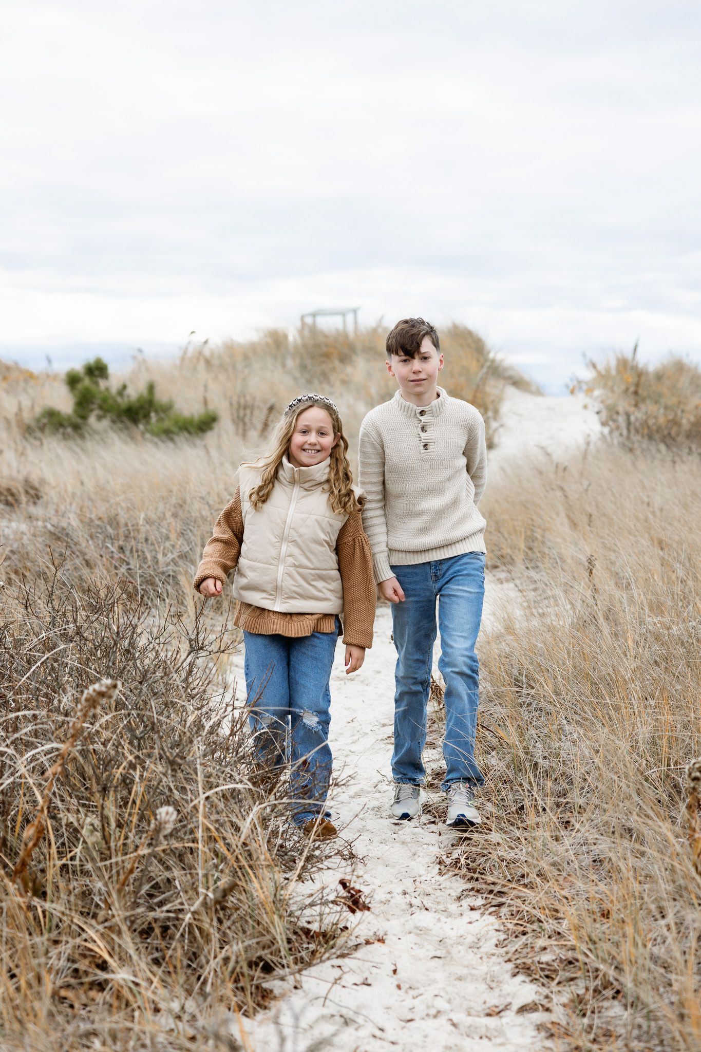 Family walking hand in hand along sandy path through coastal grass.