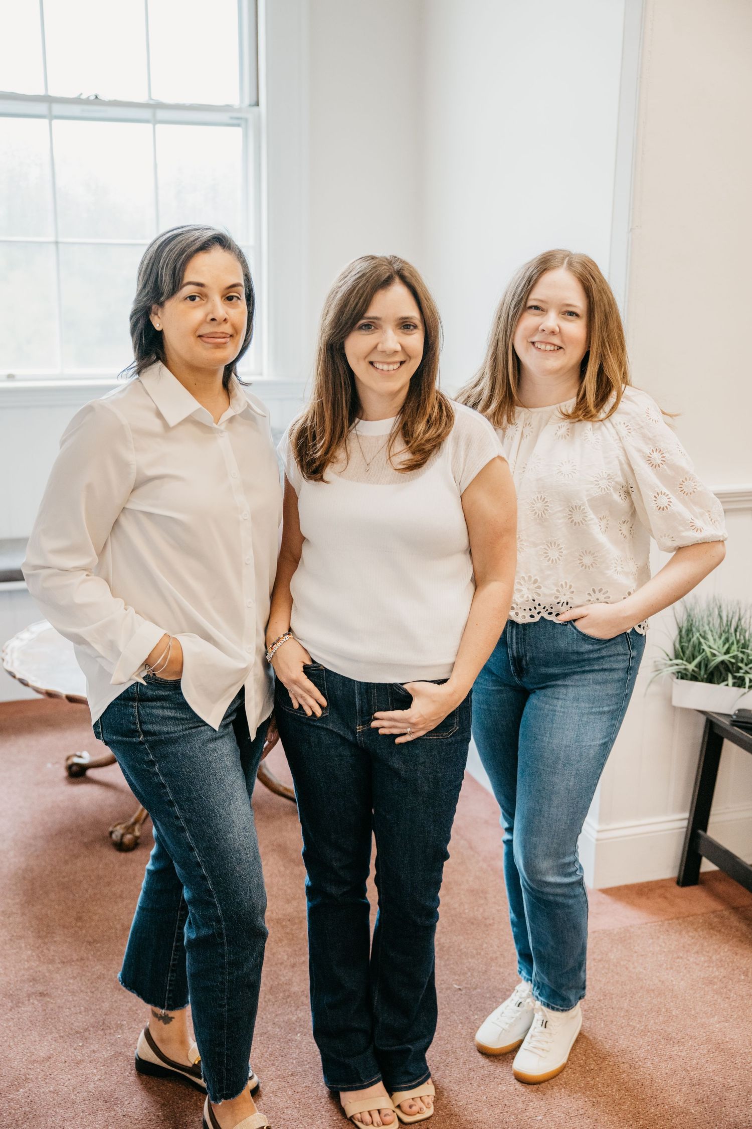 Three women in white tops and jeans smile together indoors in a bright, modern office setting.