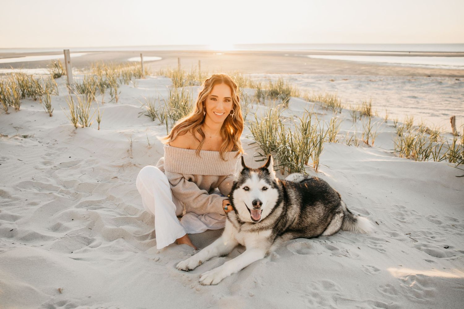 Woman smiling with Siberian Husky on sandy beach at golden sunset hour.