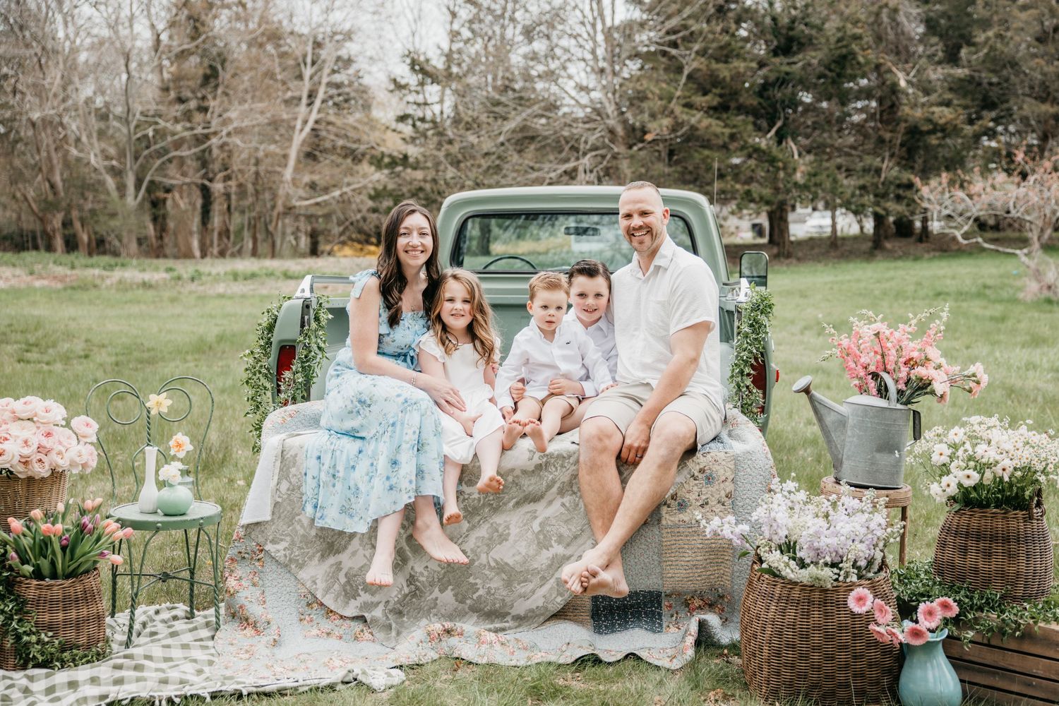 Family of five posing on vintage green truck bed surrounded by spring flowers in lush green outdoor setting.
