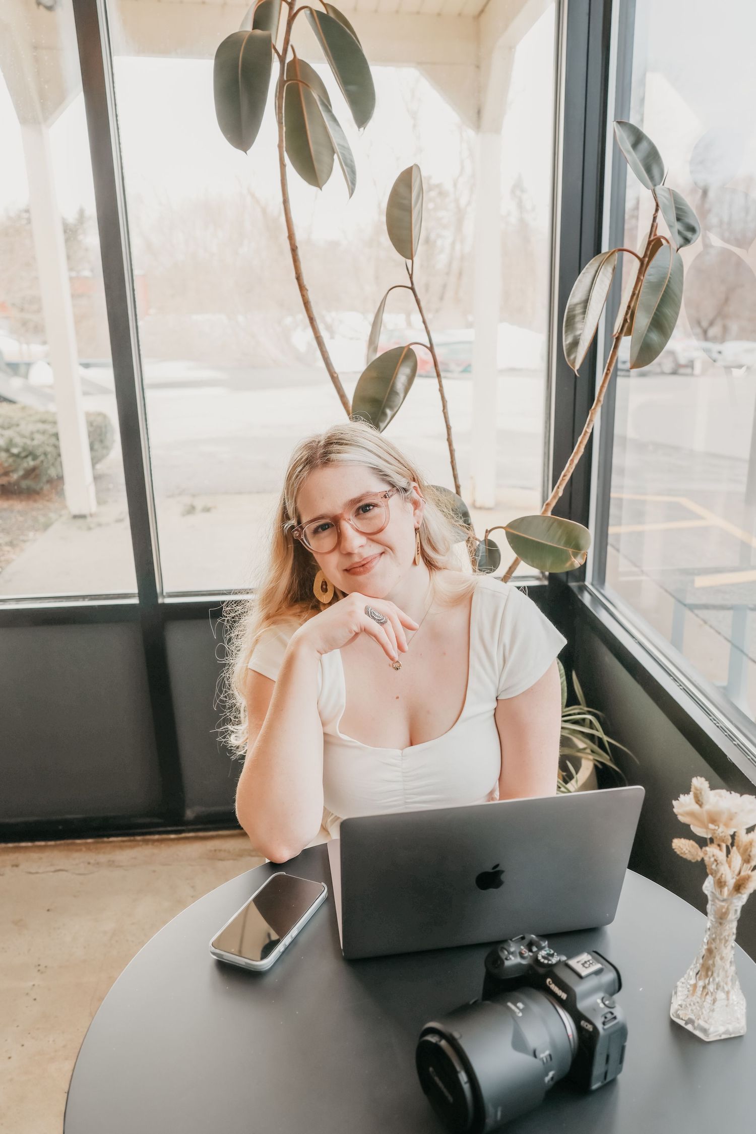 A person sits at a desk with a laptop, houseplant, and camera in a bright sunlit room with large windows.