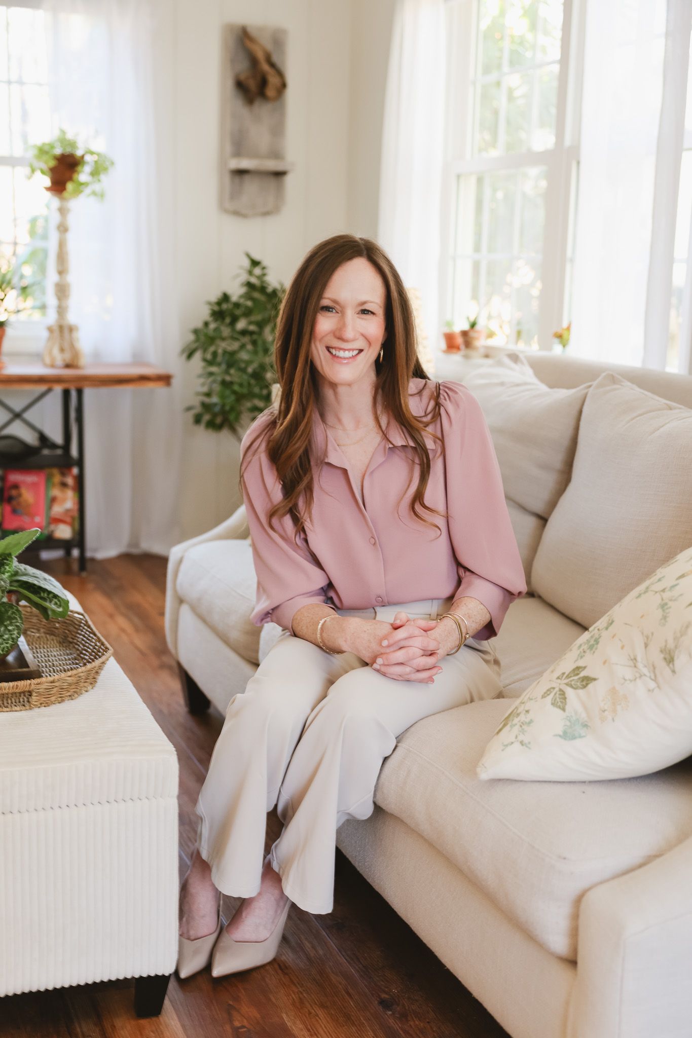 Smiling woman in pink blouse and beige pants sitting on cream sofa in bright, plant-filled living room.