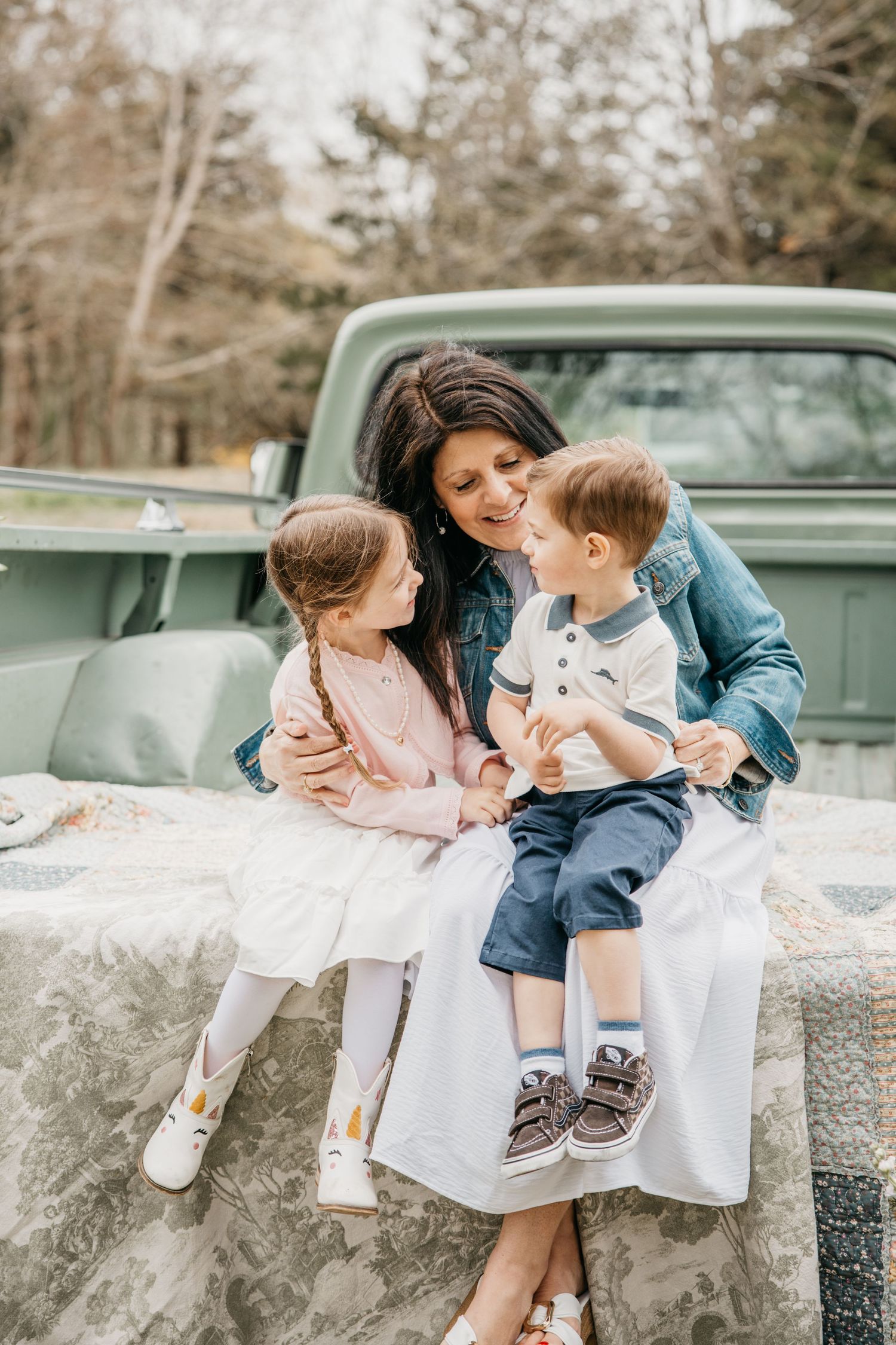 A mother sits on a vintage truck tailgate hugging her two young children outdoors near trees.