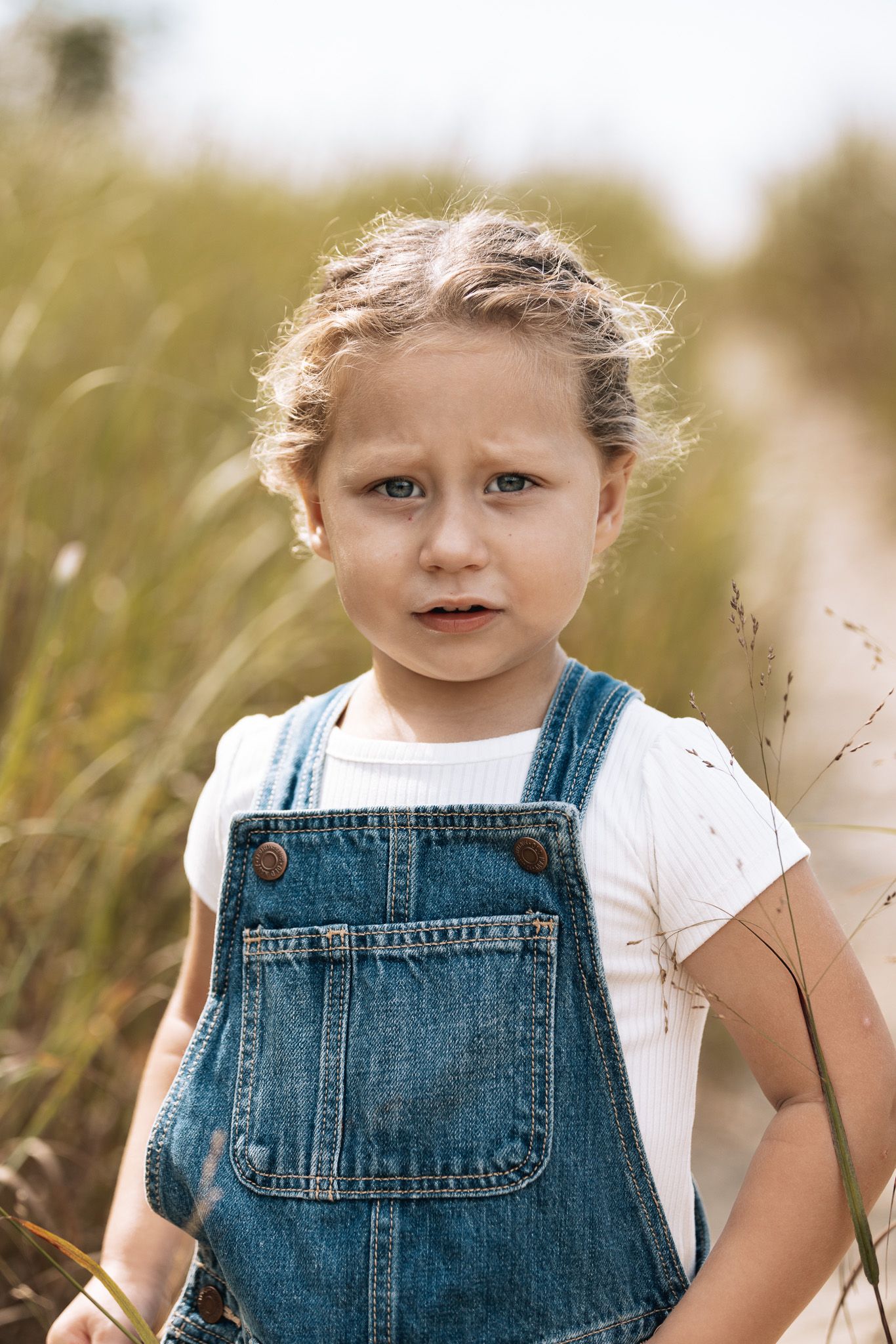 Child in denim overalls smiling while playing in tall summer grass.