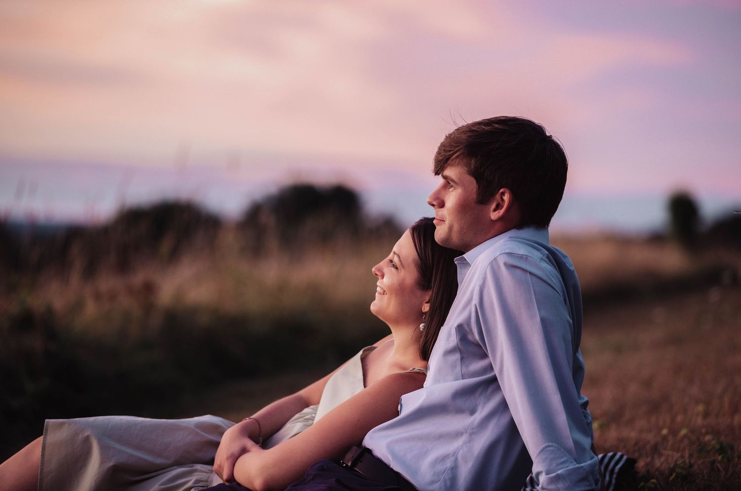 A couple embraces while watching a vibrant sunset in a peaceful field.