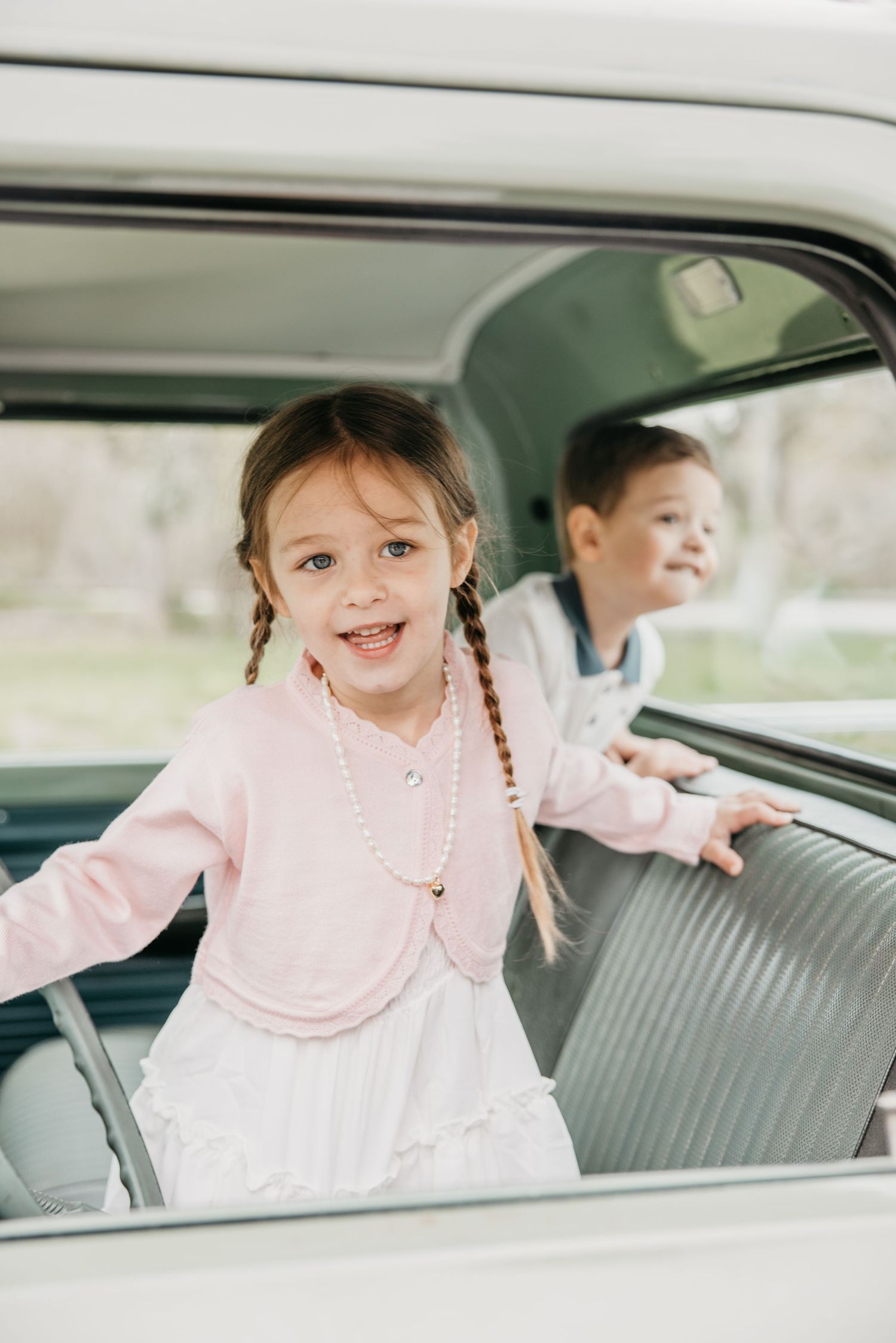 Two cheerful children in vintage car interior, girl in pink cardigan with braids and pearl necklace smiling at camera.