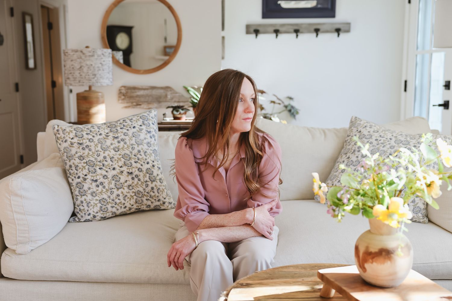 Woman in pink blouse sitting on white sofa with floral pillows in bright cozy living room with mirror and vase of flowers.