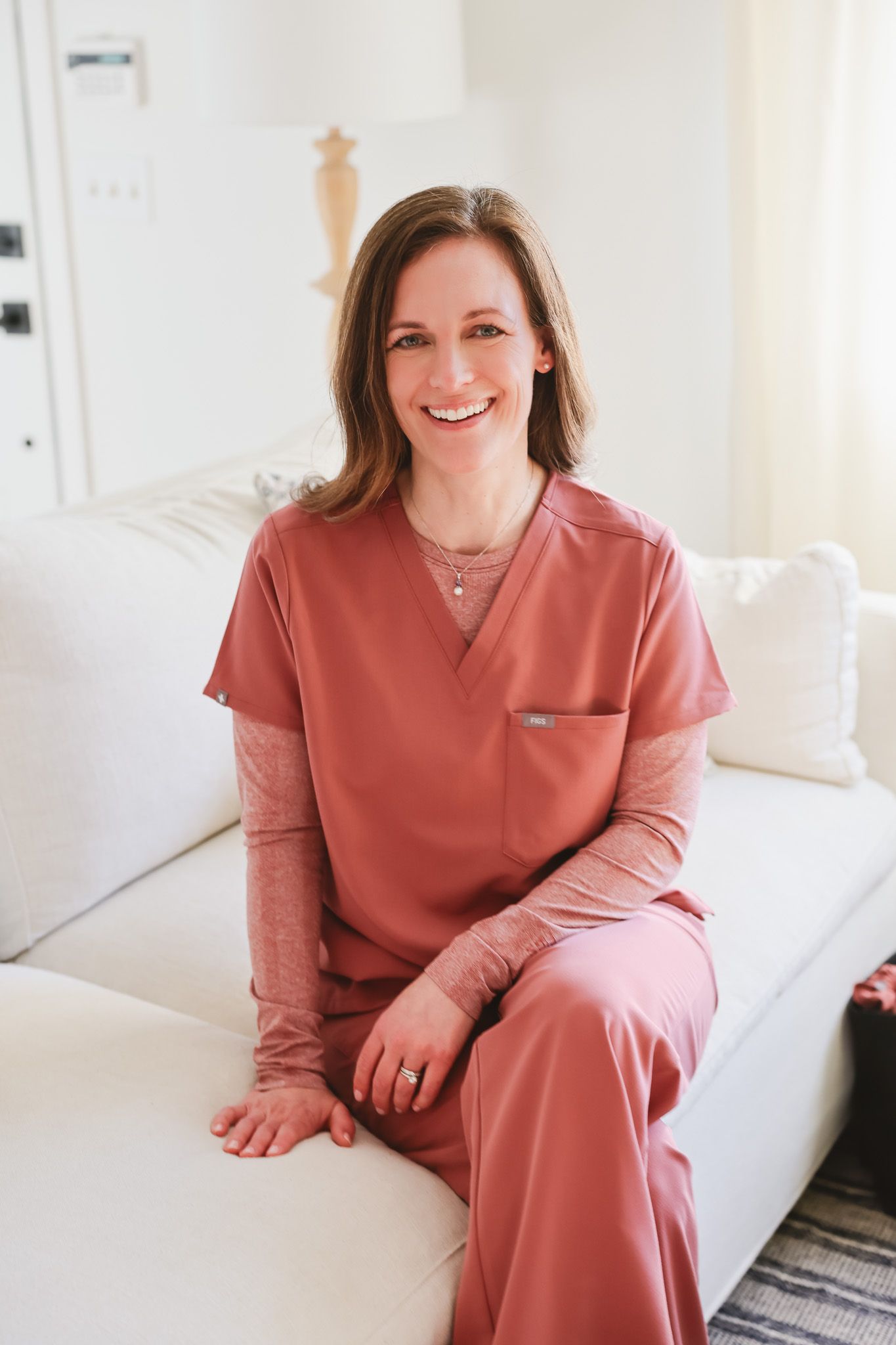 Smiling female healthcare professional in pink scrubs sitting on a white couch in a bright, modern medical office.