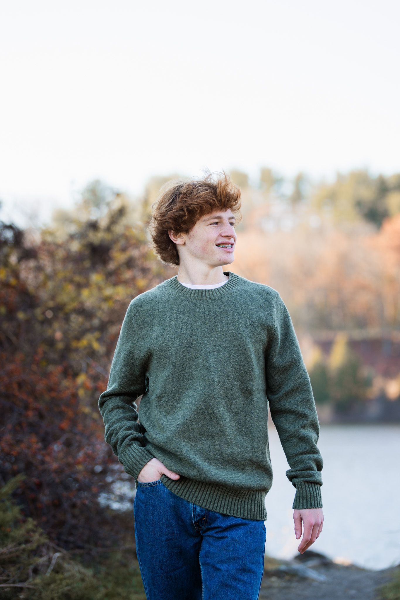 Person wearing a green sweater and blue jeans poses casually against an autumn outdoor backdrop.