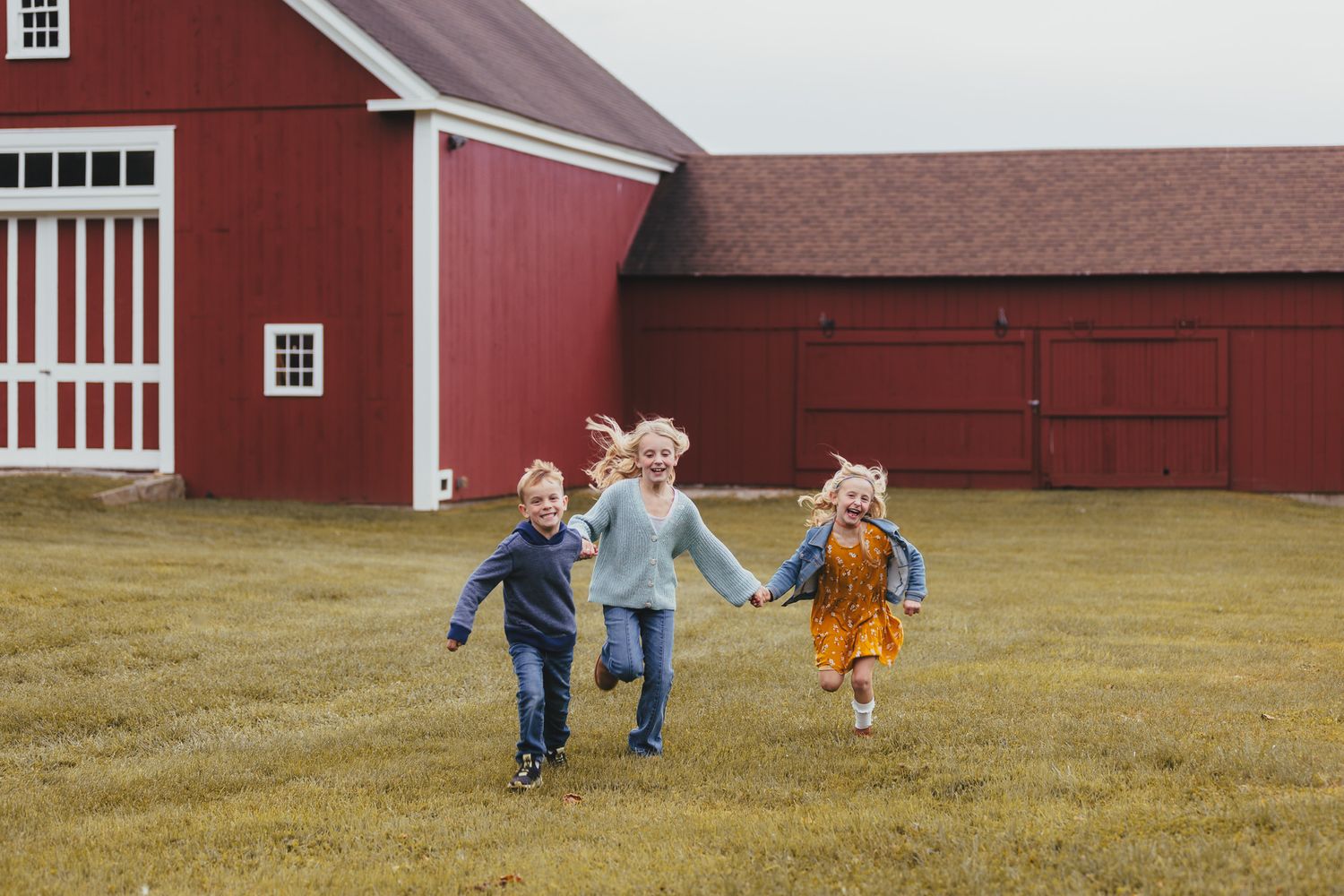 Three children running and holding hands in front of a red barn.