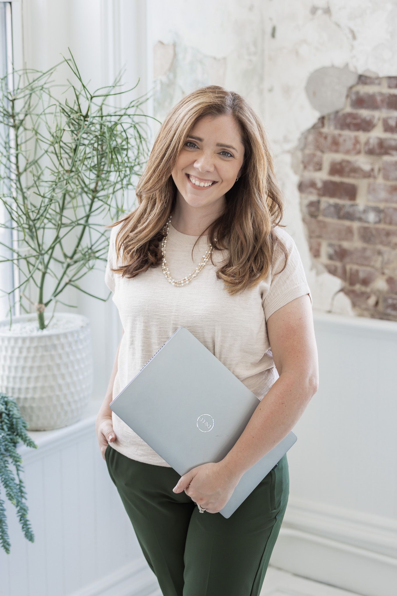 Professional wearing a white blouse and dark pants holds a laptop in a bright modern office setting.