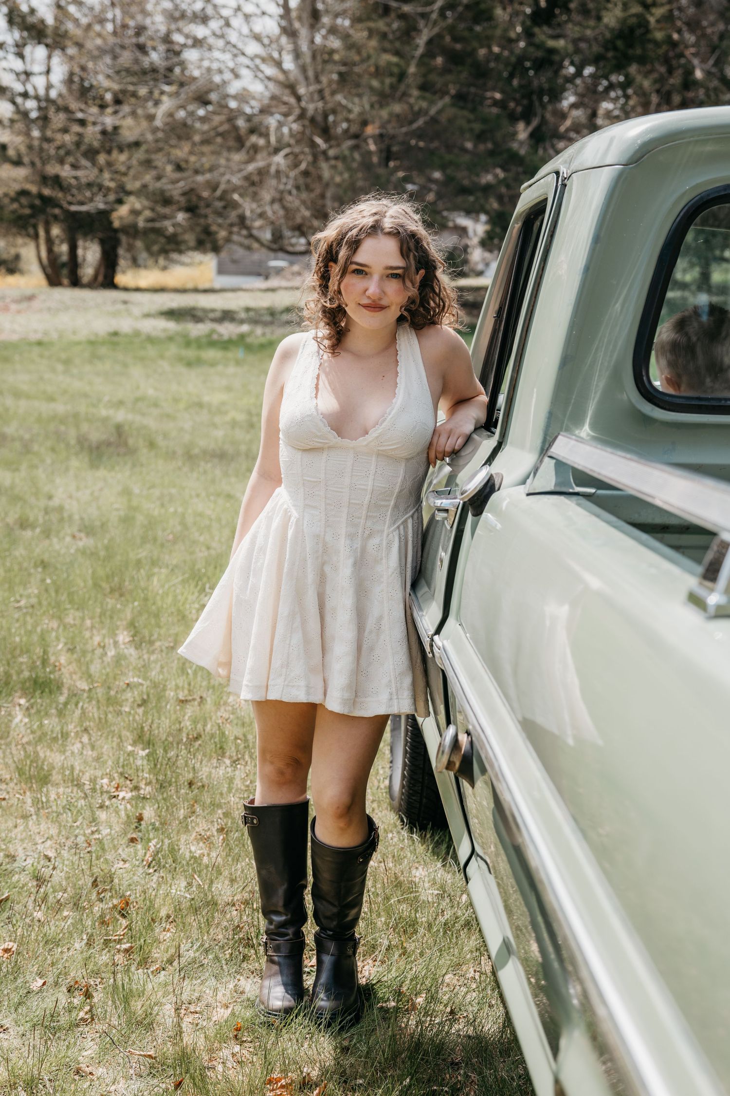 Young woman in white mini dress and black boots posing beside a vintage mint green truck on a grassy lawn.