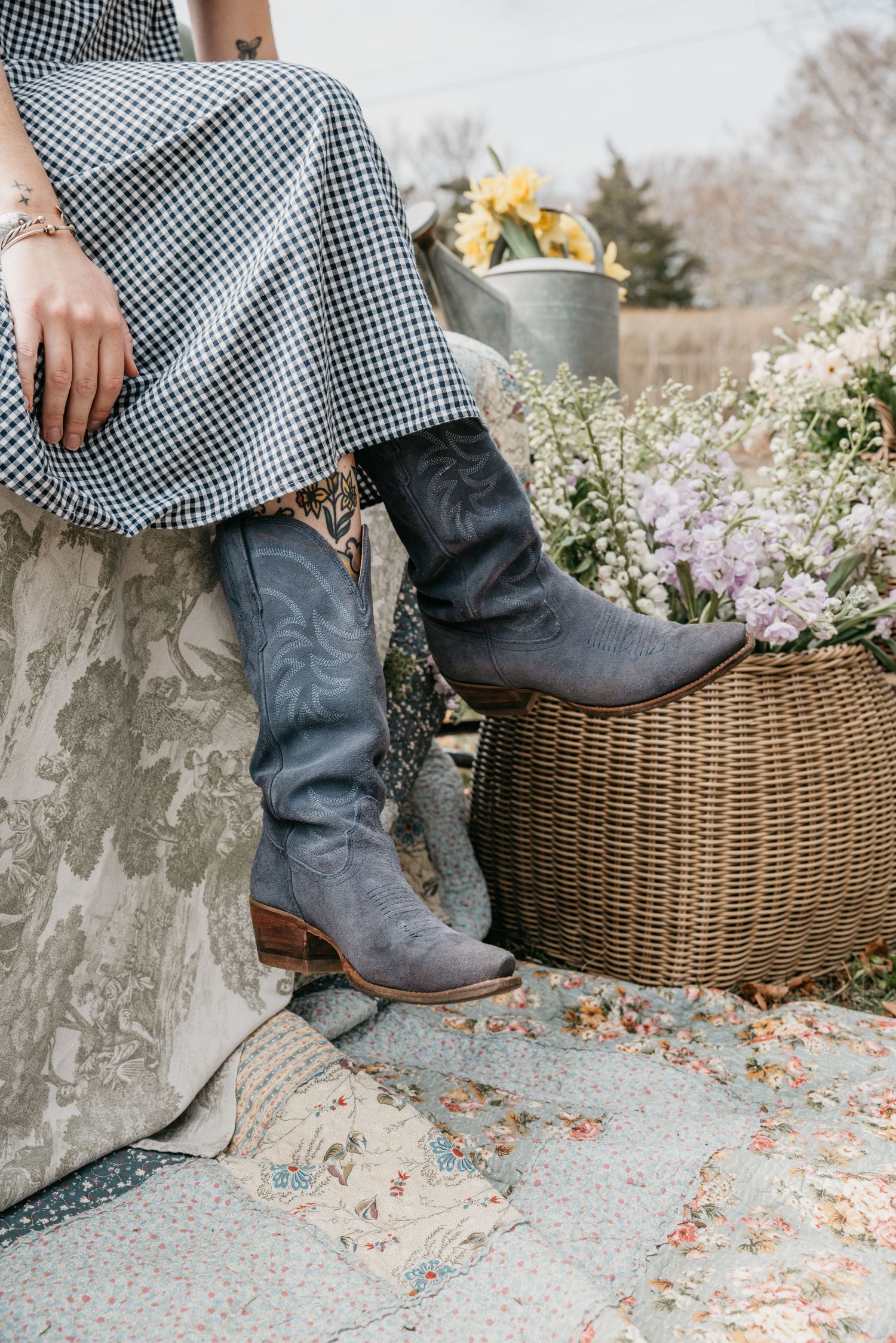 Woman in gingham dress wearing gray cowboy boots seated near a wicker basket with spring flowers.