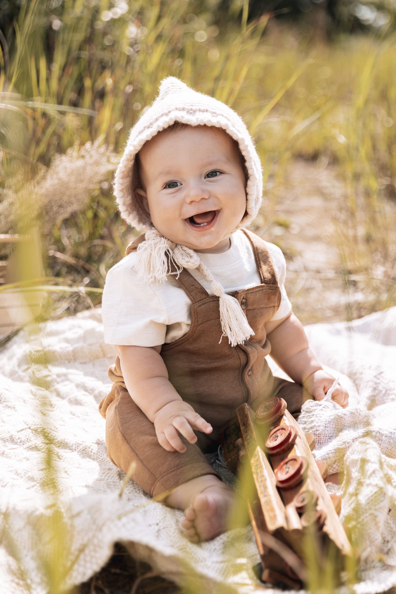 Baby wearing brown overalls and bonnet sits happily in sunlit grass.