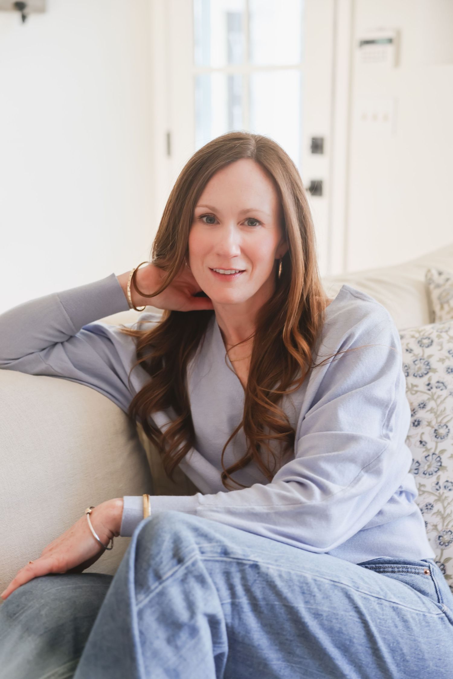 A smiling woman with long brown hair sits relaxed on a floral sofa in a bright, airy room wearing a blue outfit.