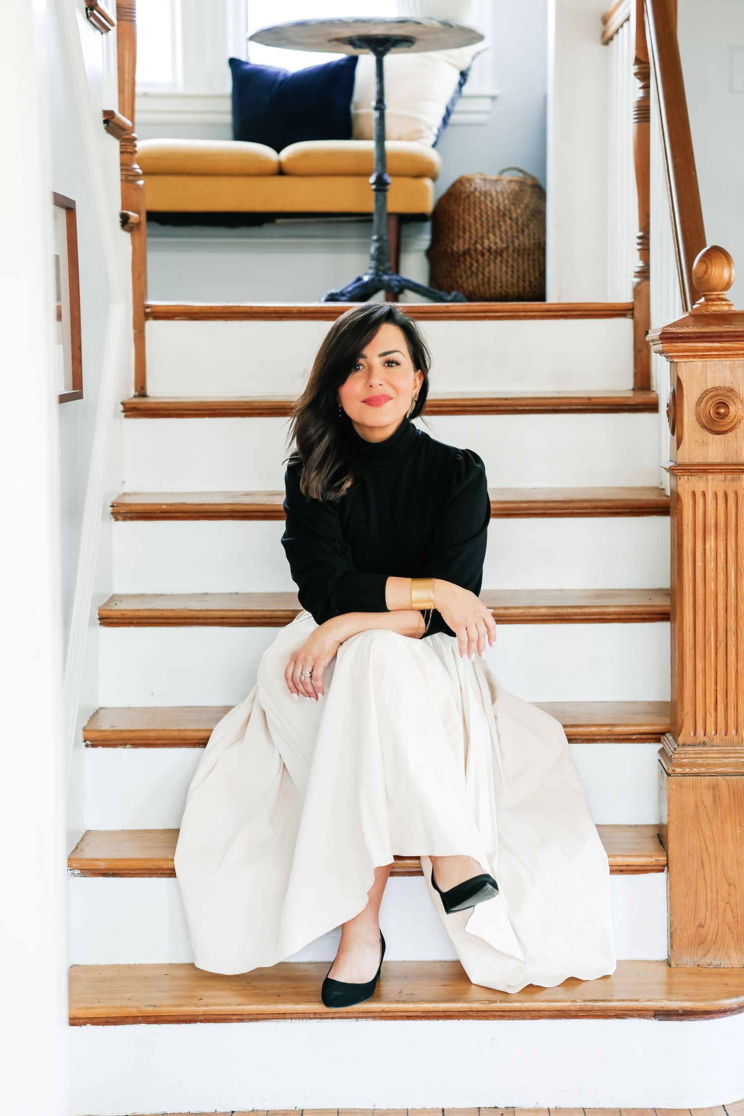 Woman in black turtleneck and white wide-leg pants sitting on wooden staircase in elegant home interior.