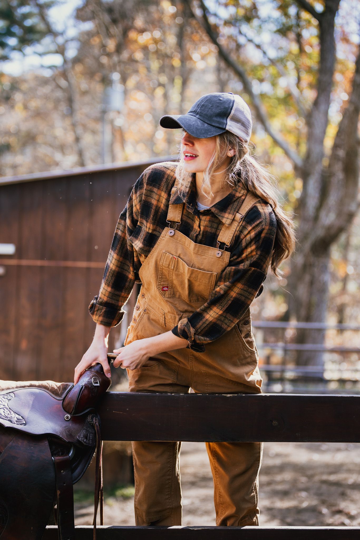 Person in brown overalls and plaid shirt leaning against wooden railing in natural setting.