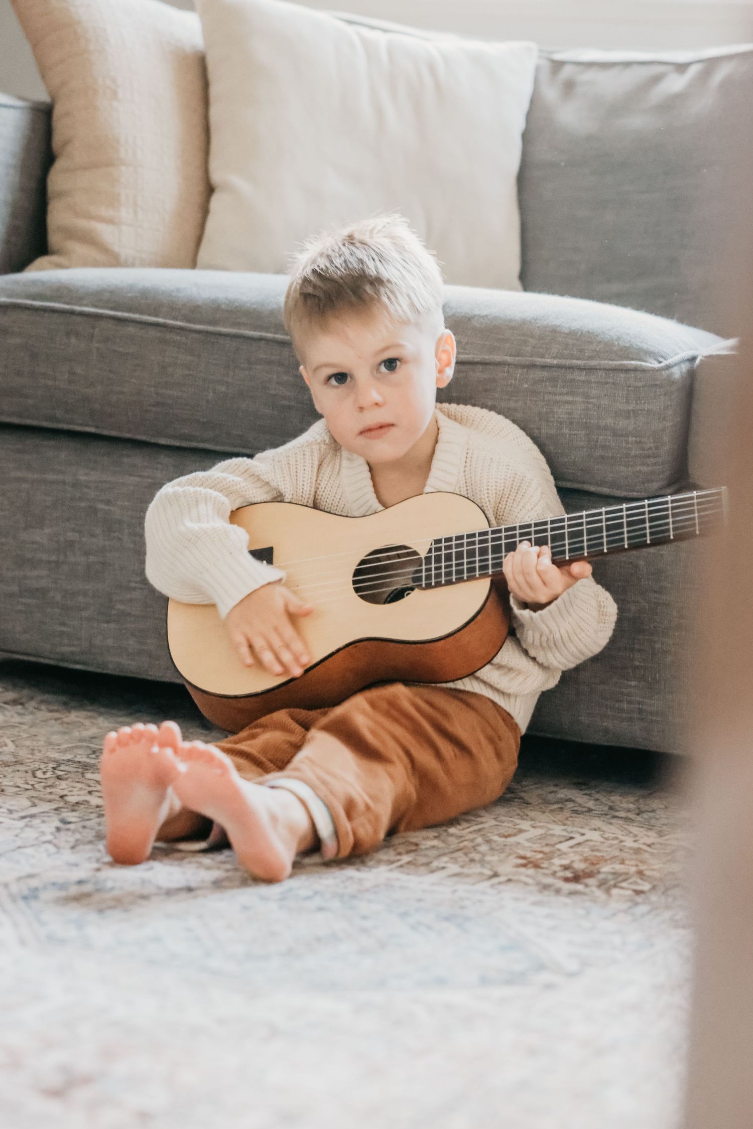 A small child sits barefoot on a carpet playing with a wooden ukulele near a gray couch.