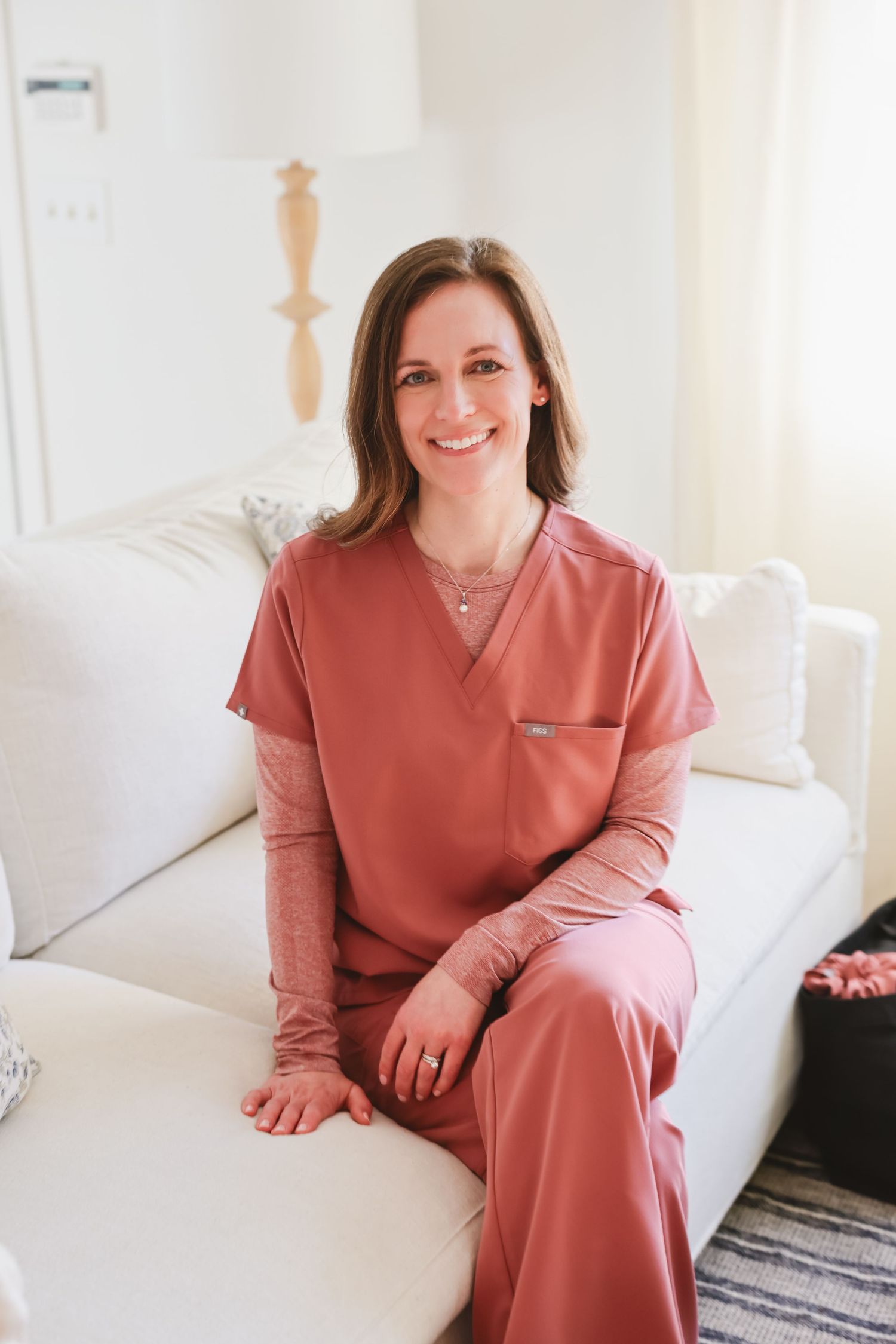 Smiling woman in pink scrubs sitting on a white couch in a bright, modern room.