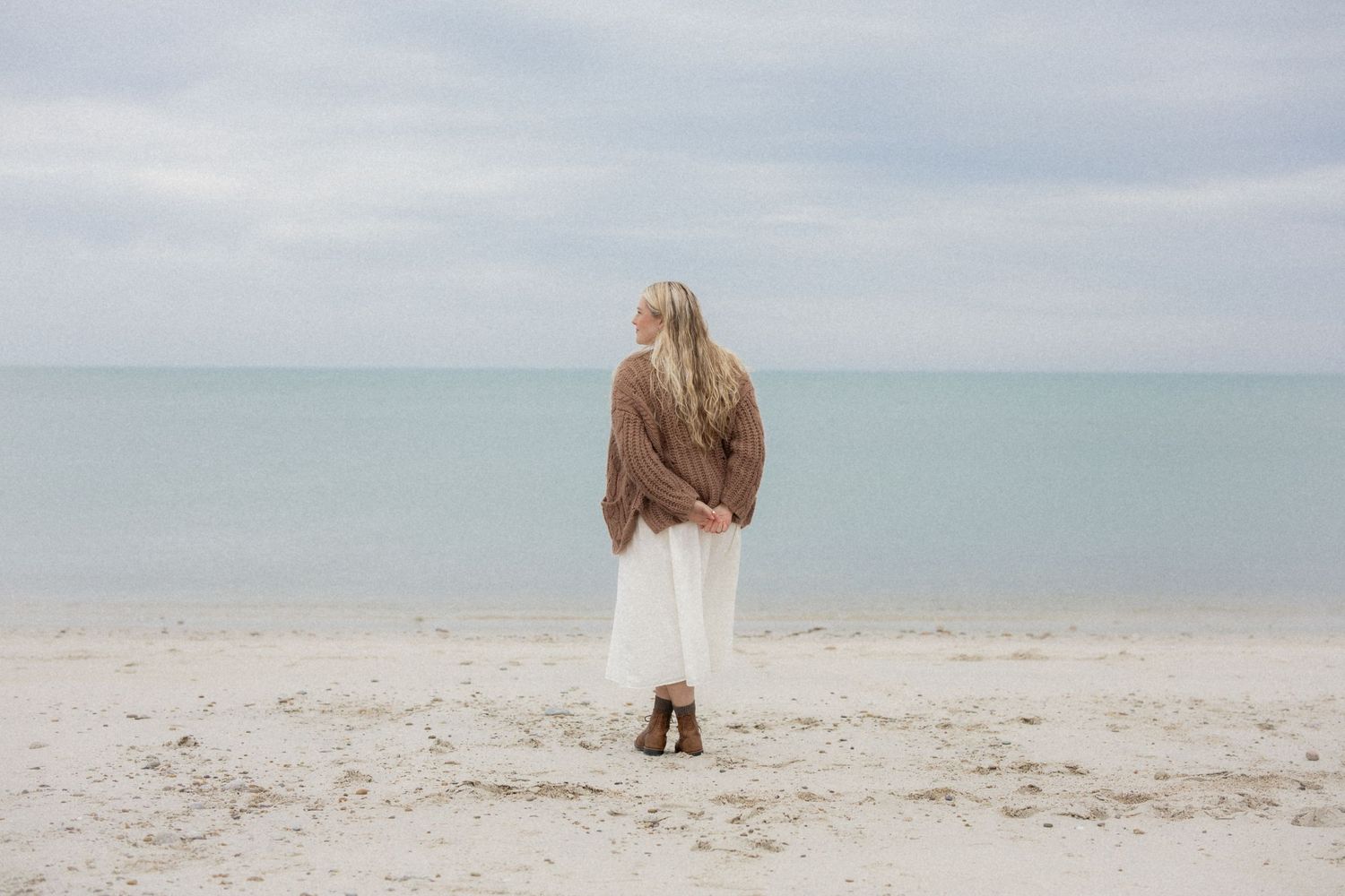 Person in brown sweater and white skirt stands alone on a misty beach looking out at the calm ocean.