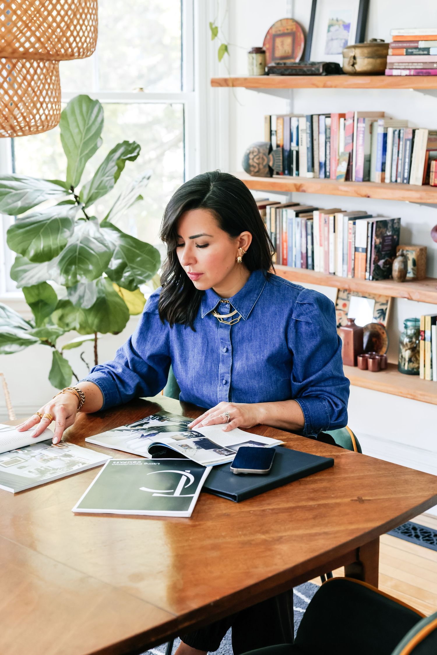 Woman in blue shirt reviews photos and documents at wooden desk with bookshelves and large plant in bright home office.