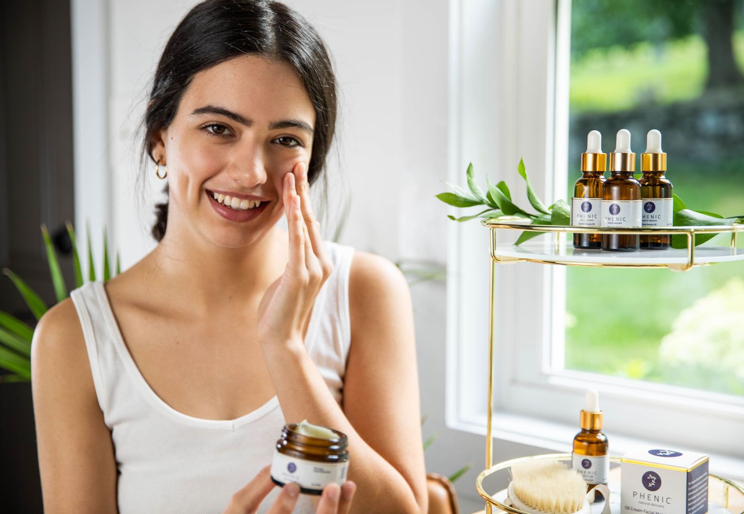 Person applying natural skincare product near window with essential oils display.