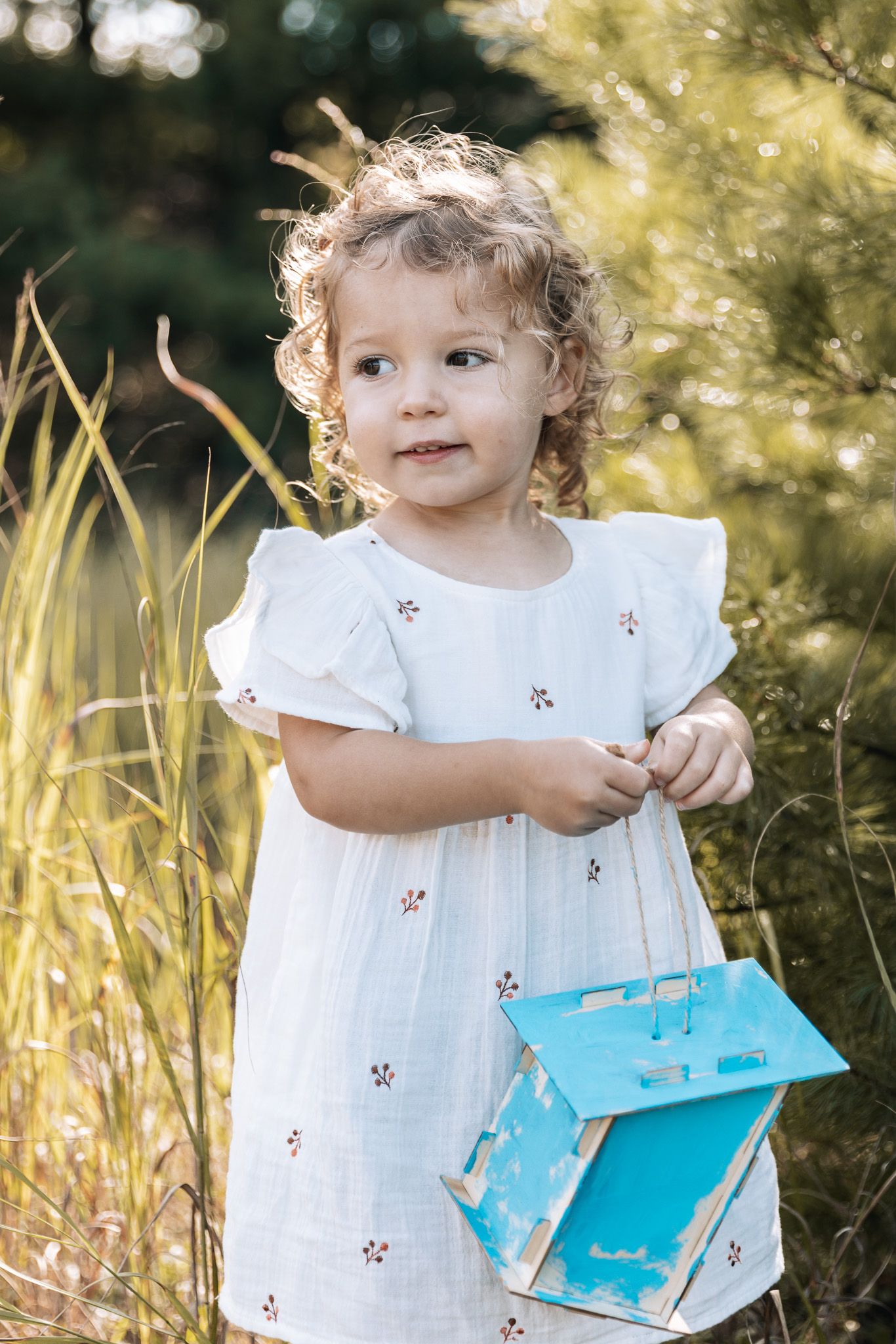 A child holds a turquoise book while wearing a white eyelet dress outdoors.