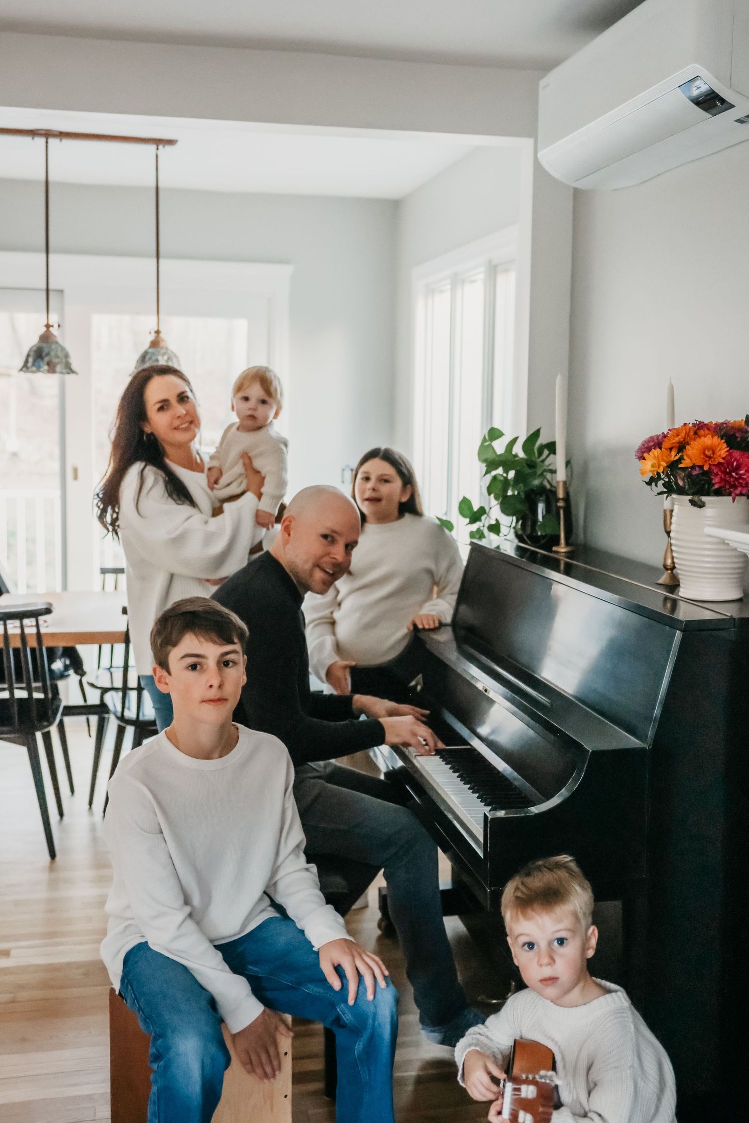A family gathering around a black piano in a bright modern living room with hardwood floors.