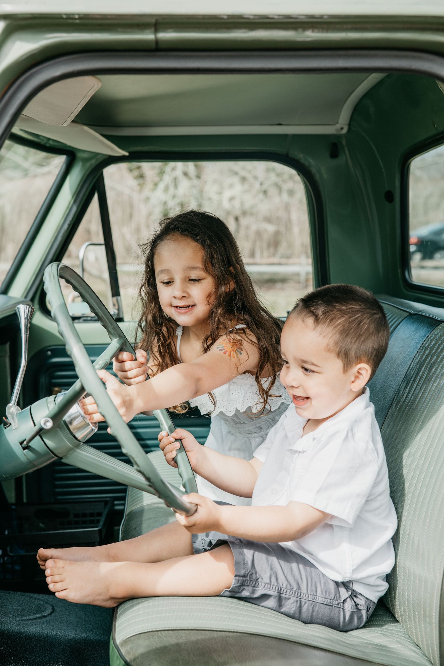 Two happy children playing with the steering wheel of a vintage green truck, sitting on a light blue bench seat.