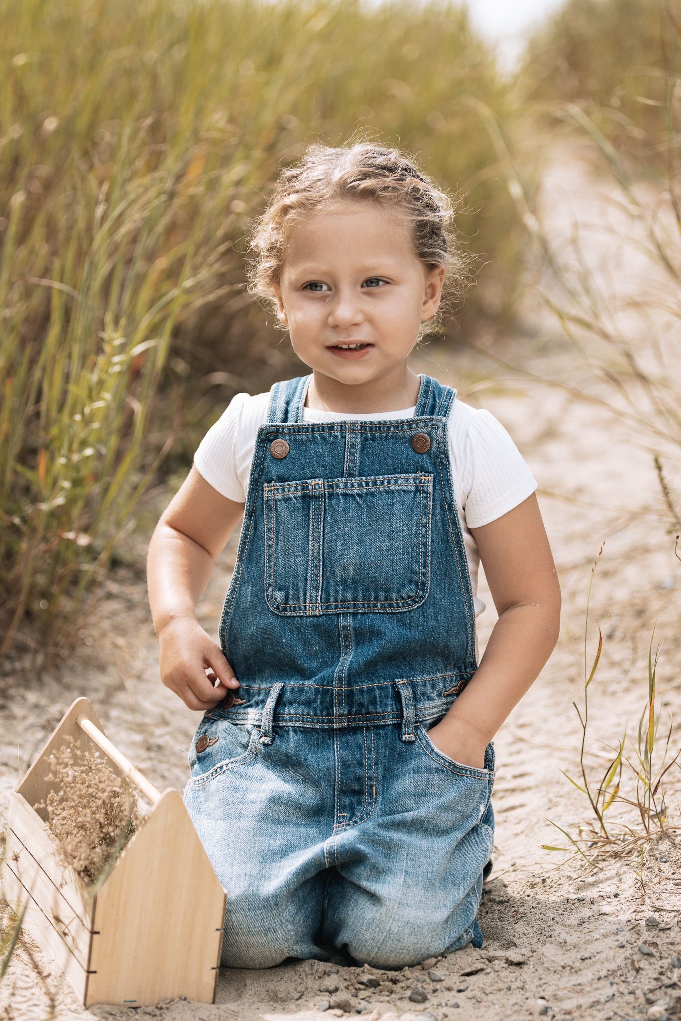 Child wearing overalls sits and plays in sandy area surrounded by coastal vegetation.